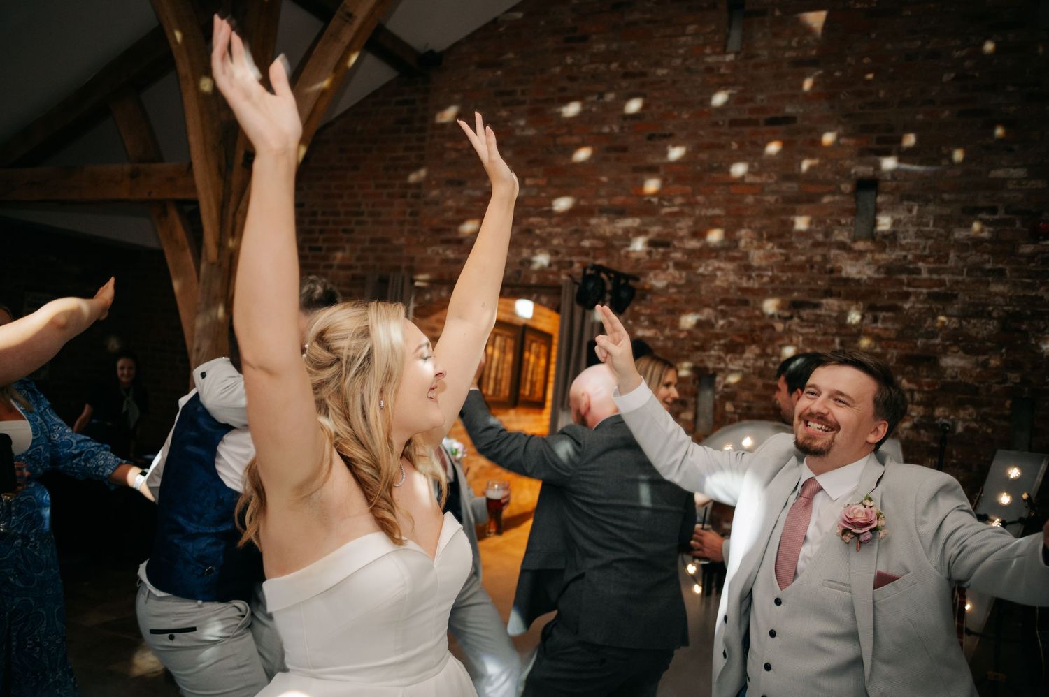 Wedding guests dancing and celebrating at a reception in a brick-walled rustic venue with festive lighting.