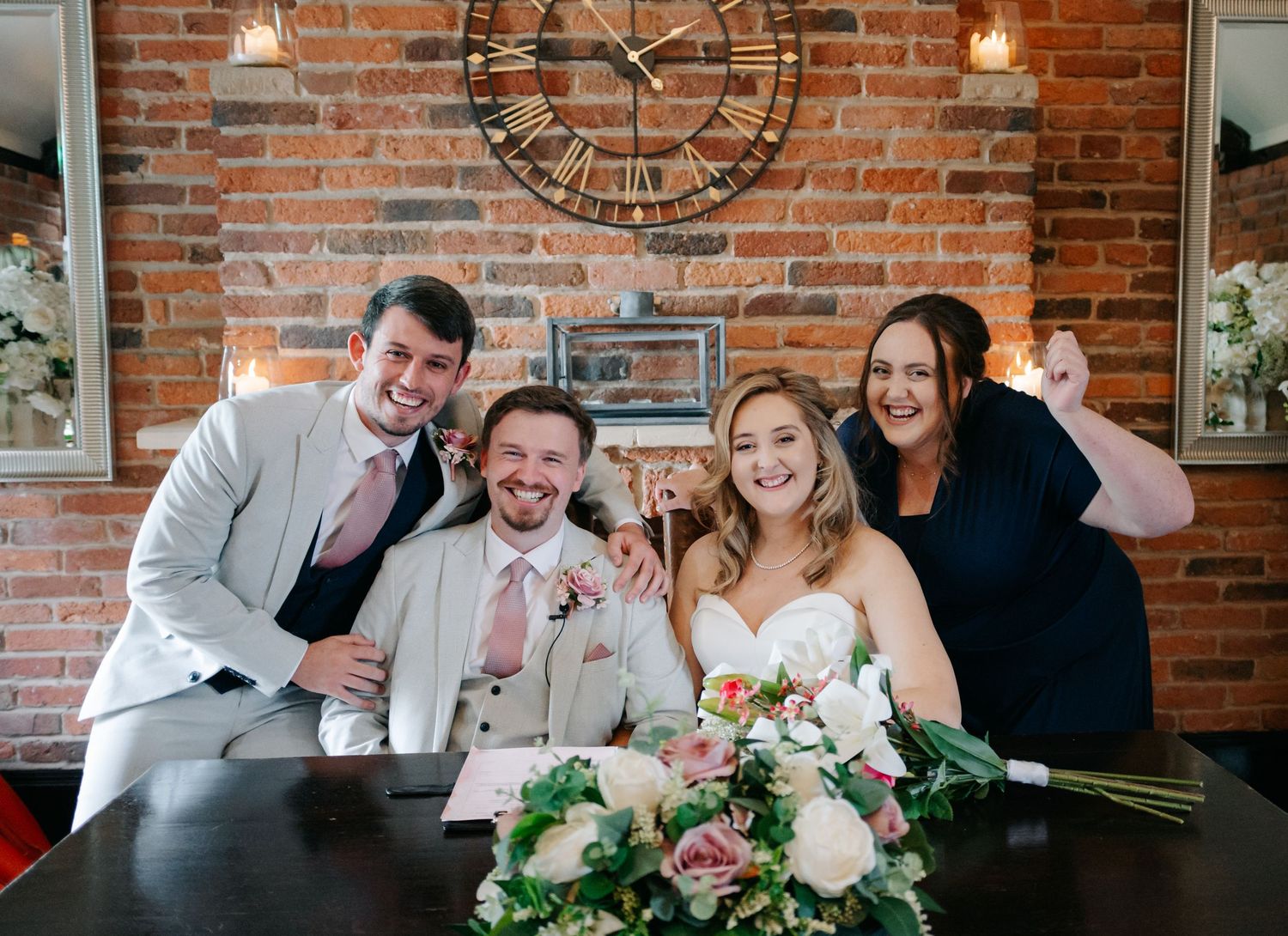 Wedding party poses for a fun group photo against a brick wall with a large clock decoration.