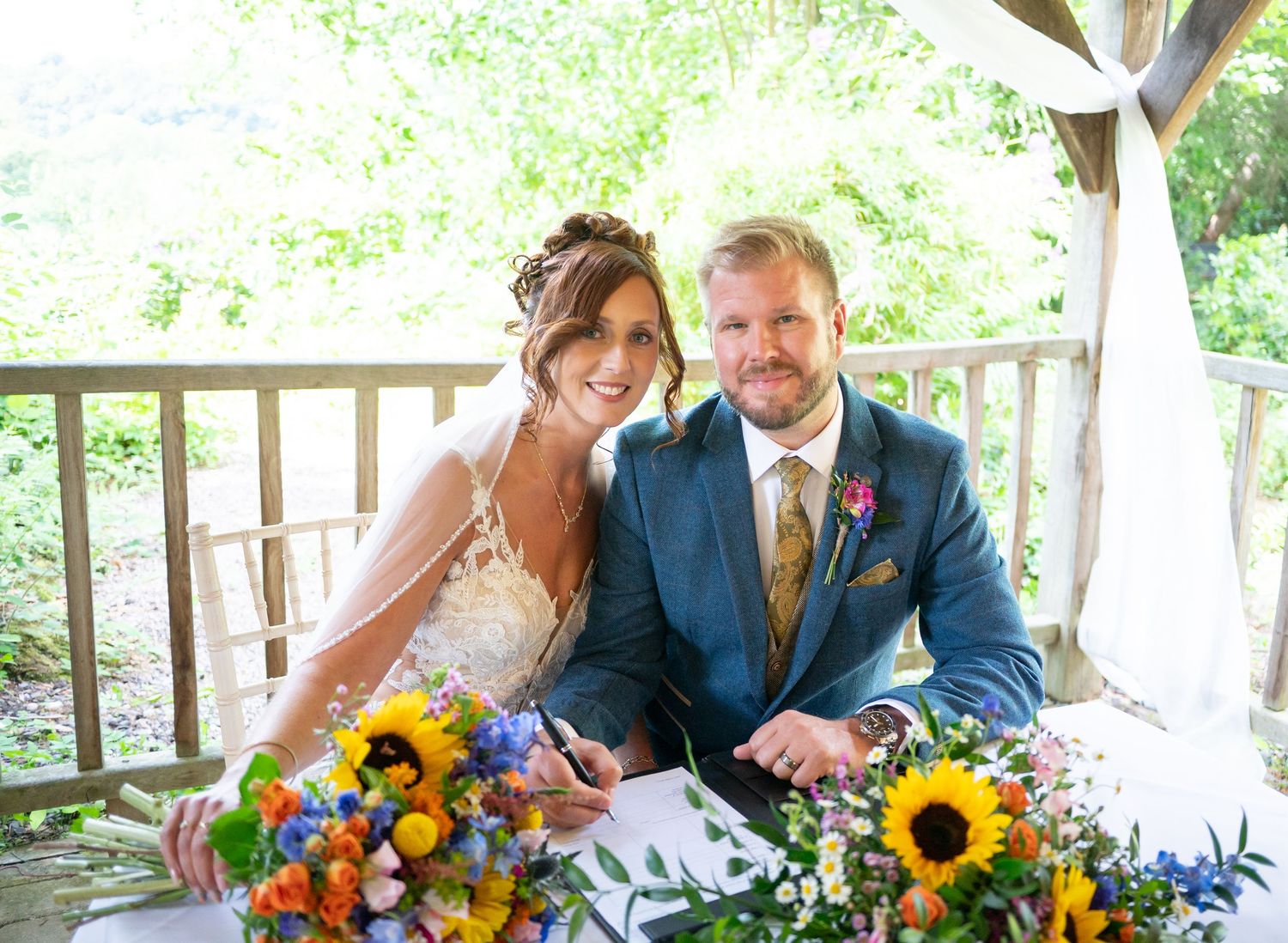 A wedding couple sits together with vibrant sunflower bouquet under a covered outdoor venue.