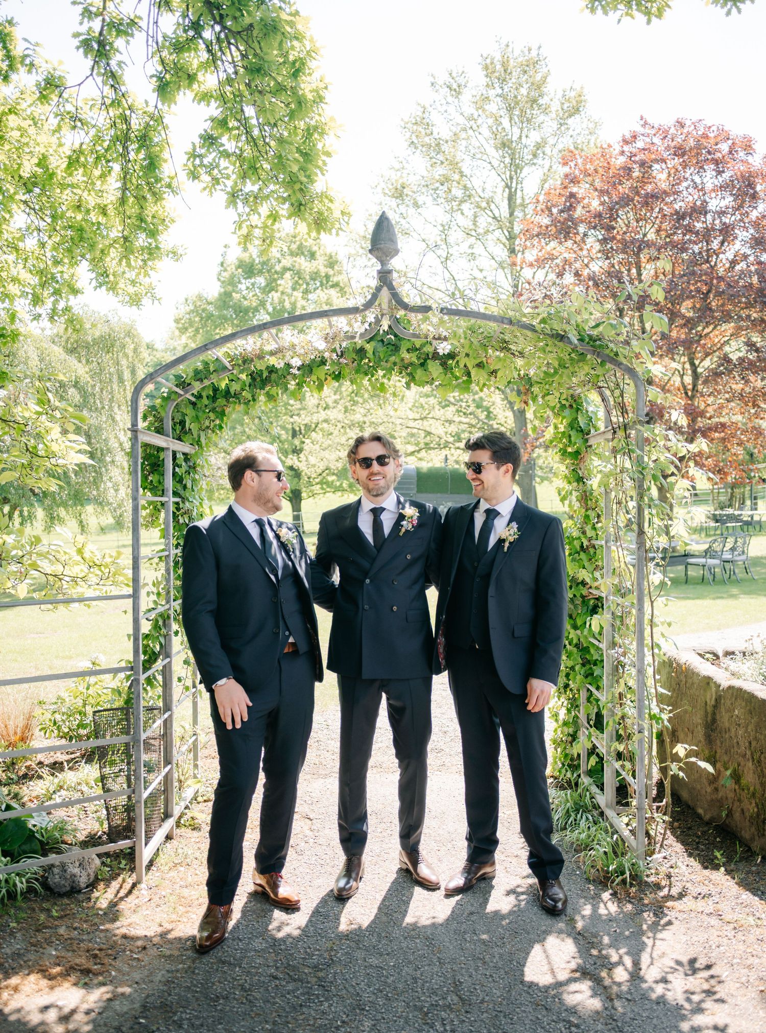 Groomsmen in black suits pose together on a stone path surrounded by lush greenery.