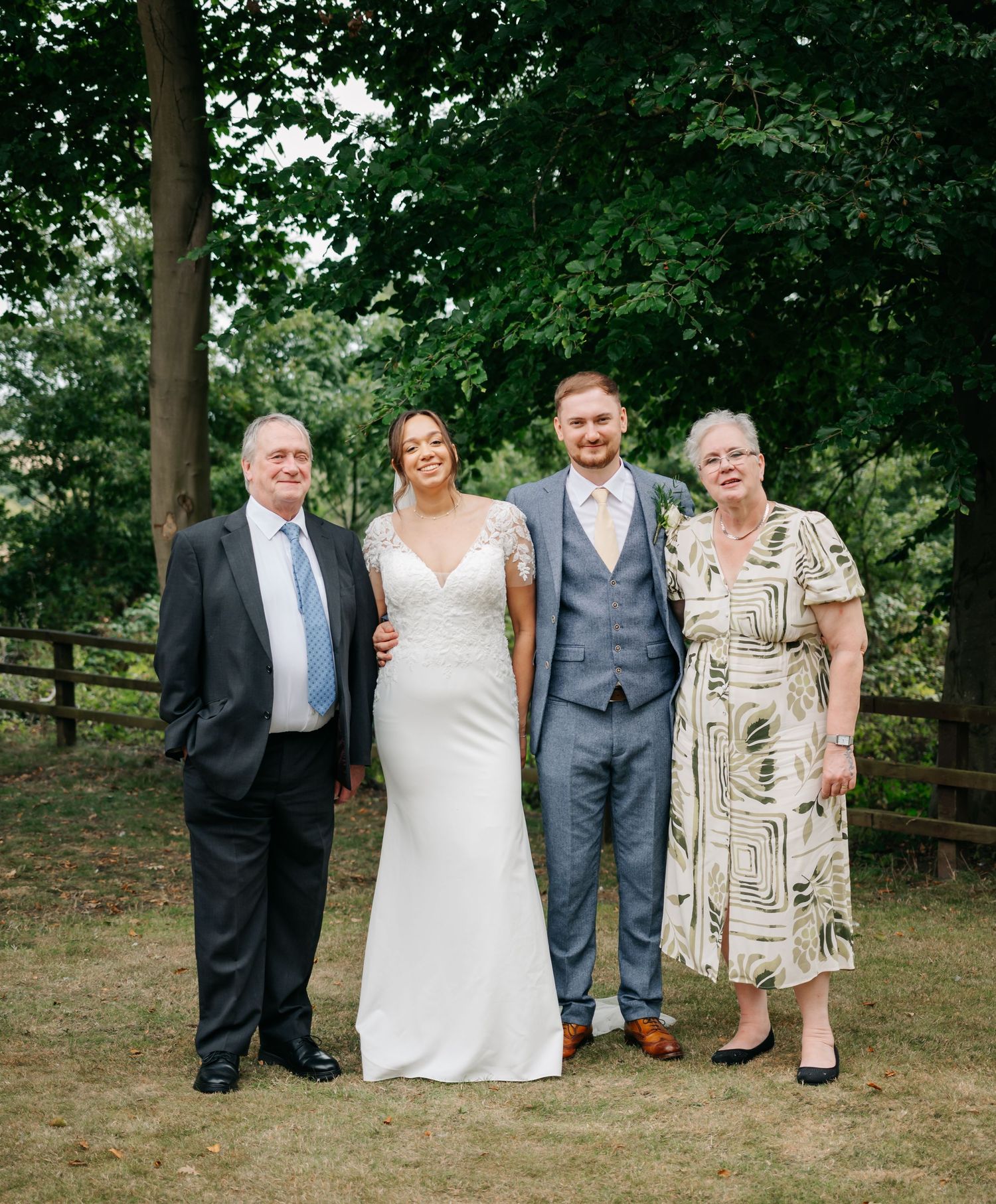 A wedding party of four people poses together outdoors in front of trees and a wooden fence.
