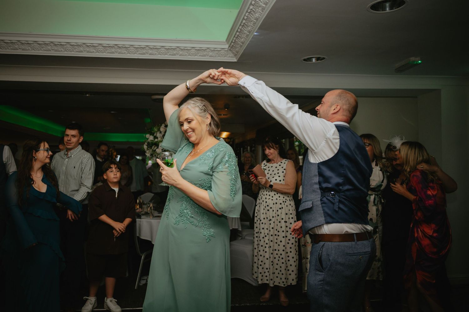 Wedding reception dance floor filled with guests celebrating under green lighting.