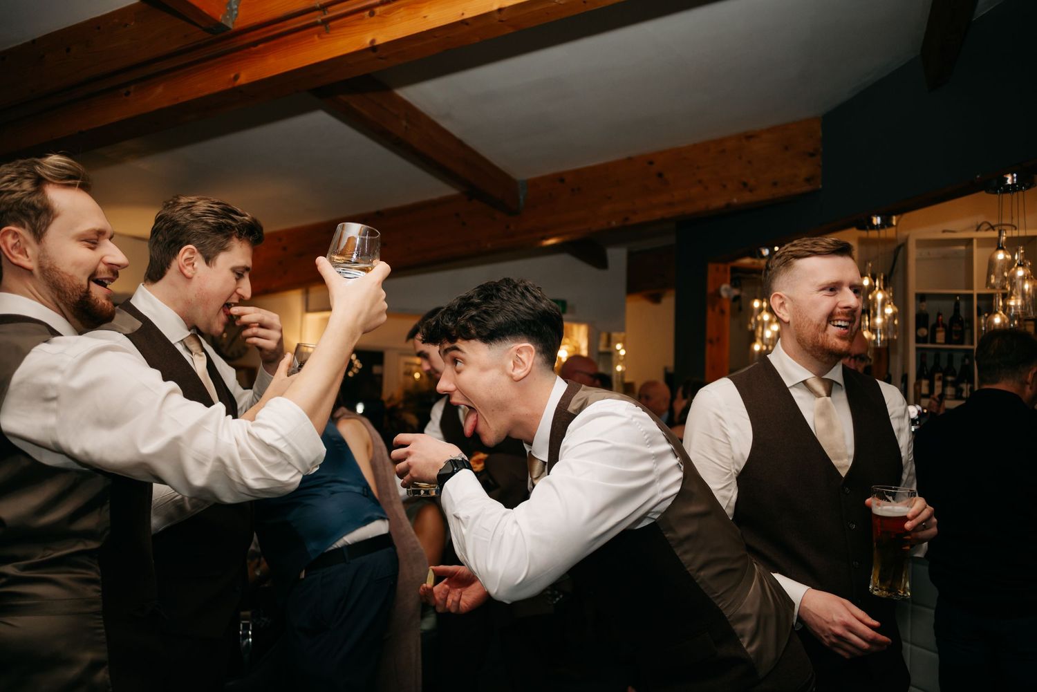 Wedding reception party with guests dancing under wooden beams in warm lighting.