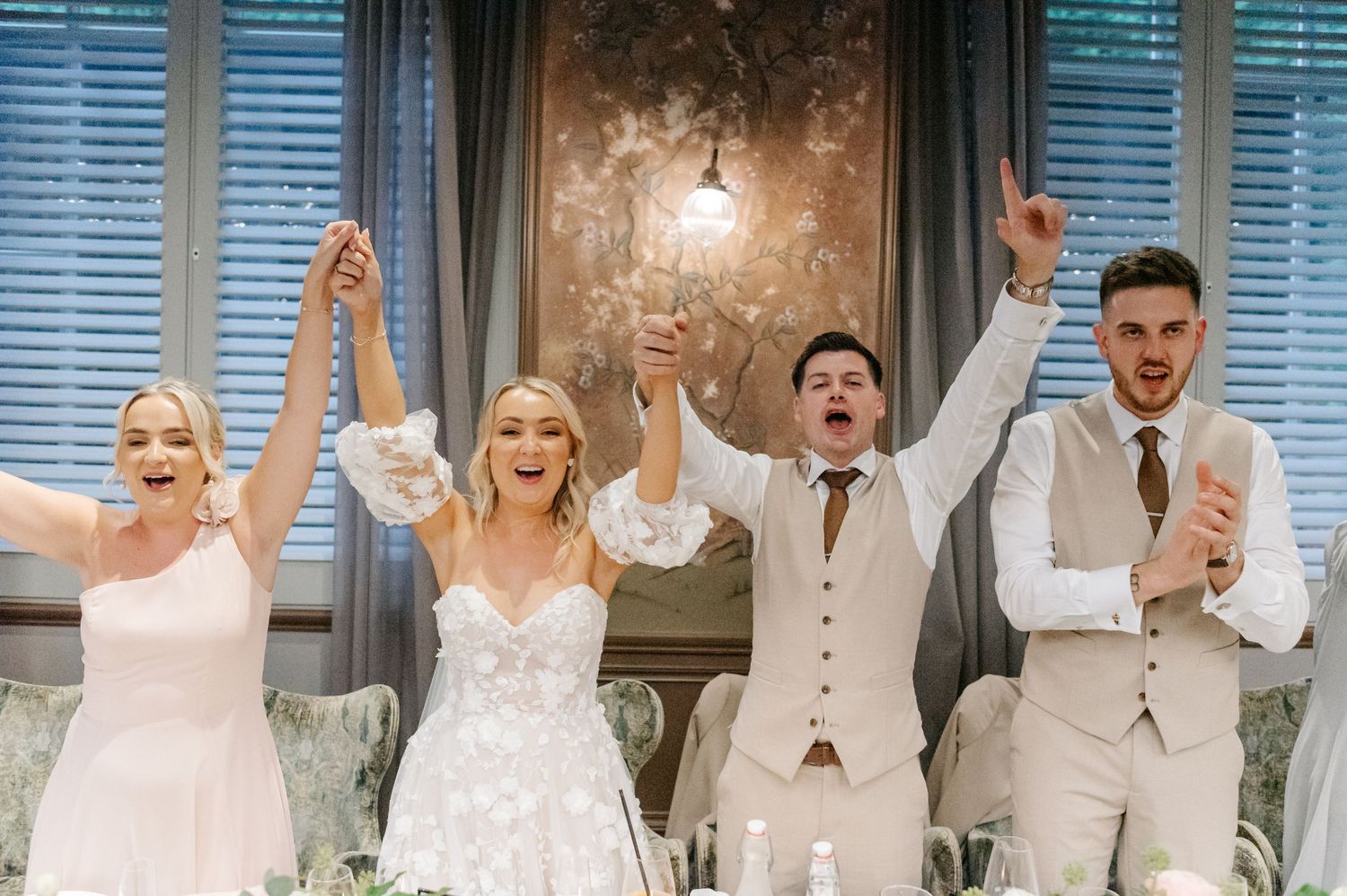 Wedding celebration group shot with people raising their arms in joy inside an elegant venue.