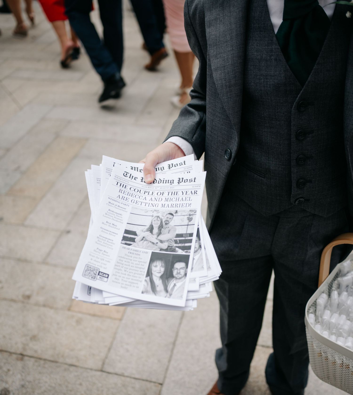 Wedding guests holding newspapers while standing on stone pavement during outdoor ceremony.