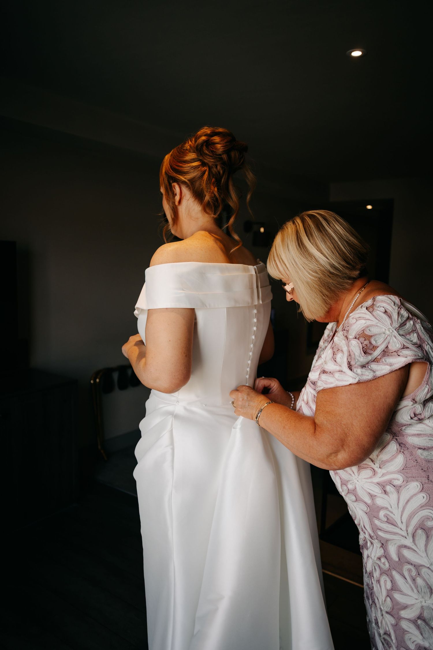 A wedding dress being laced up and adjusted from behind in a dimly lit room.