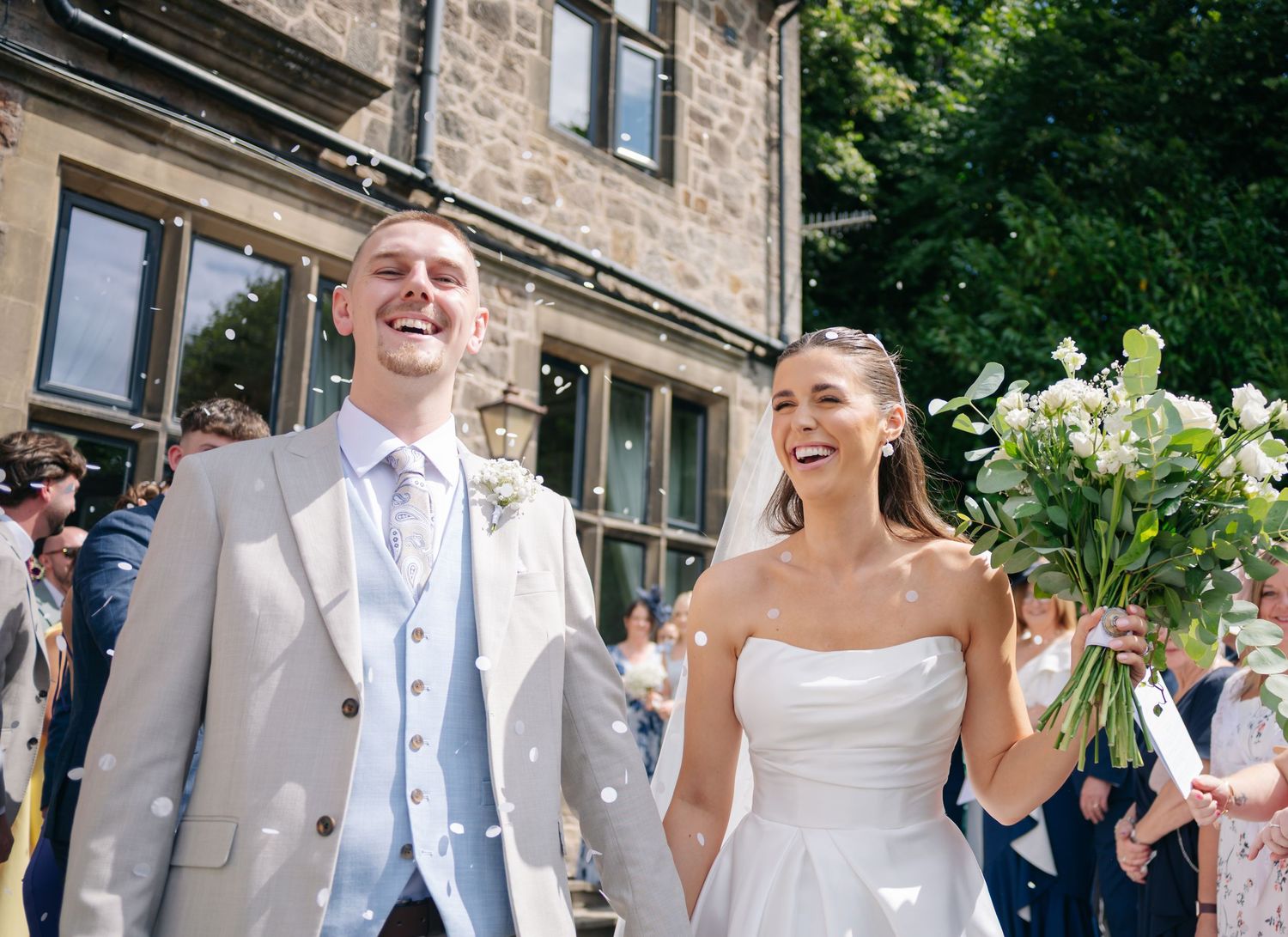 Wedding party in light gray suits and white dress walking outside historic stone building.