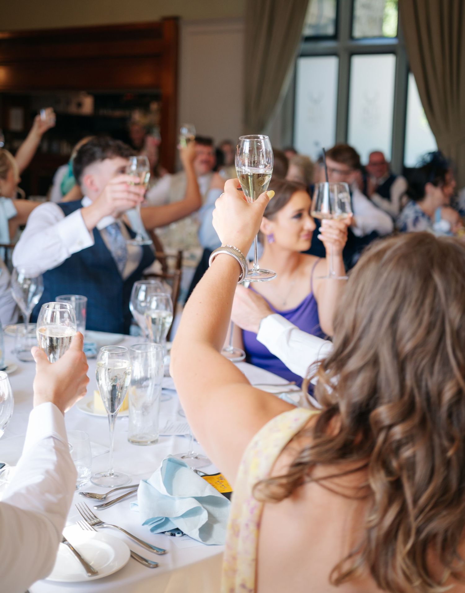 Wedding reception guests seated at round tables in a bright banquet hall with natural lighting.