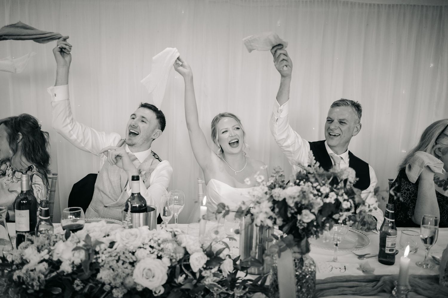 Wedding reception guests raise their glasses in celebration at a table decorated with floral arrangements.