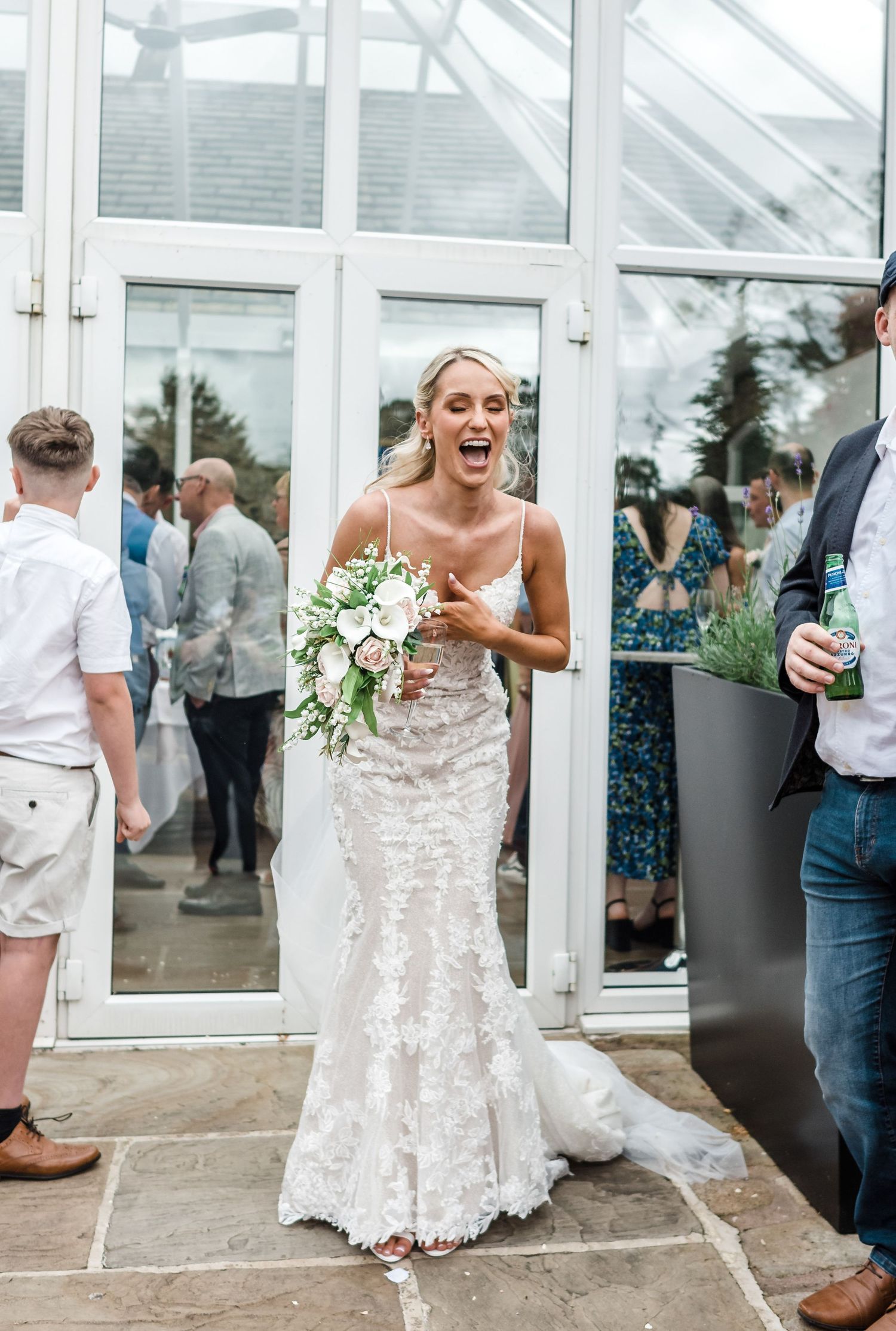 Bride in fitted lace wedding dress laughing with guests at glass conservatory venue.