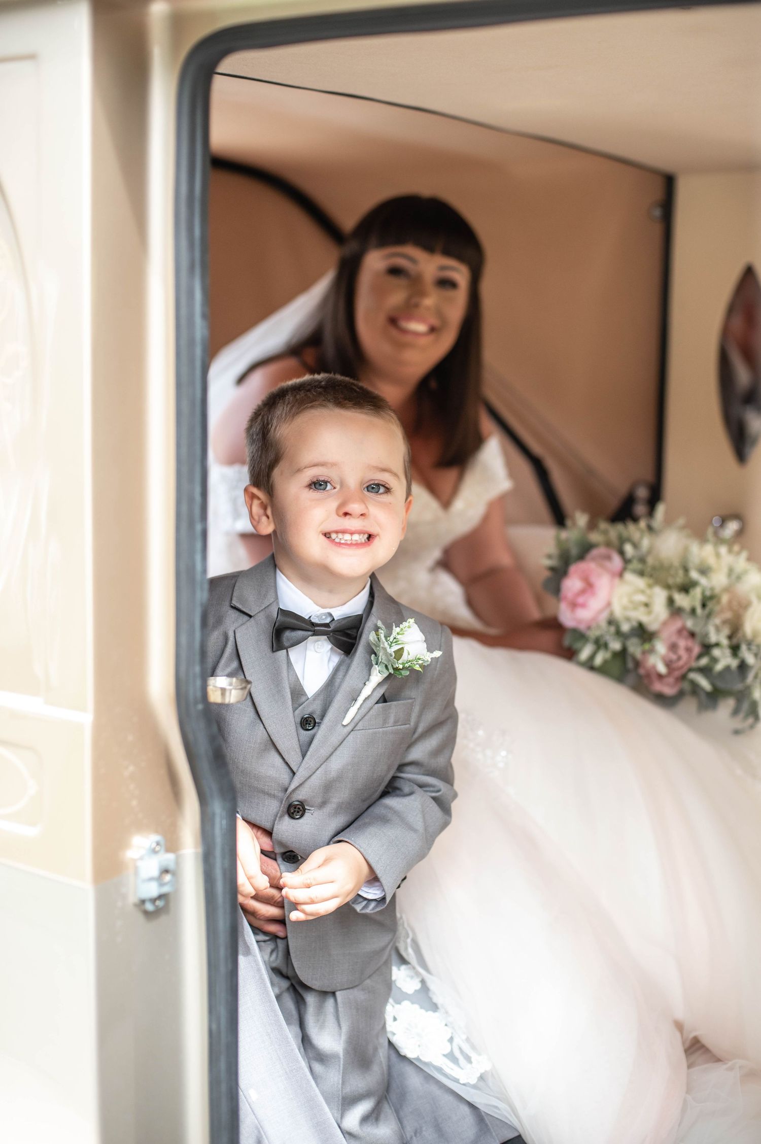A young ring bearer in a grey suit poses inside a vintage car with flowers.