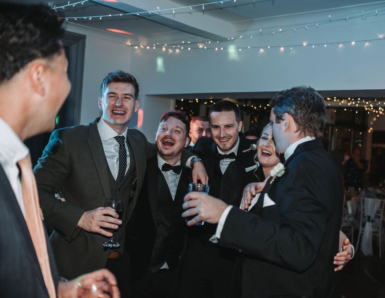 Groomsmen sharing laughs during wedding reception under string lights.