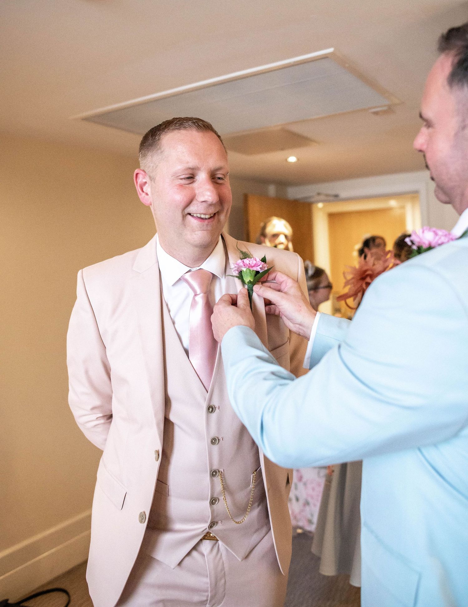 A person in a pink suit getting their boutonniere pinned by someone in a light blue jacket at formal event.