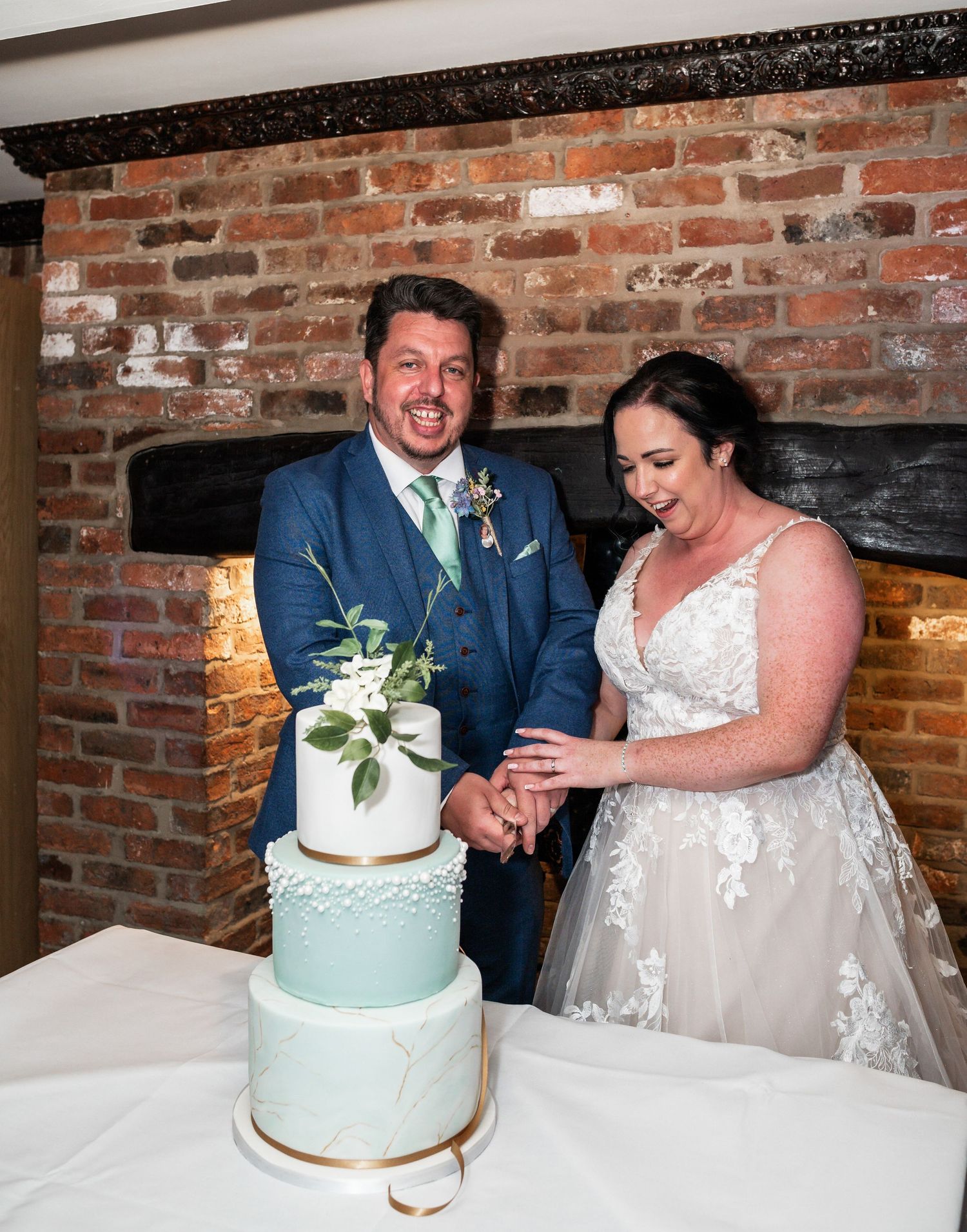 A couple cuts their three-tiered mint green and white wedding cake against a brick wall backdrop.