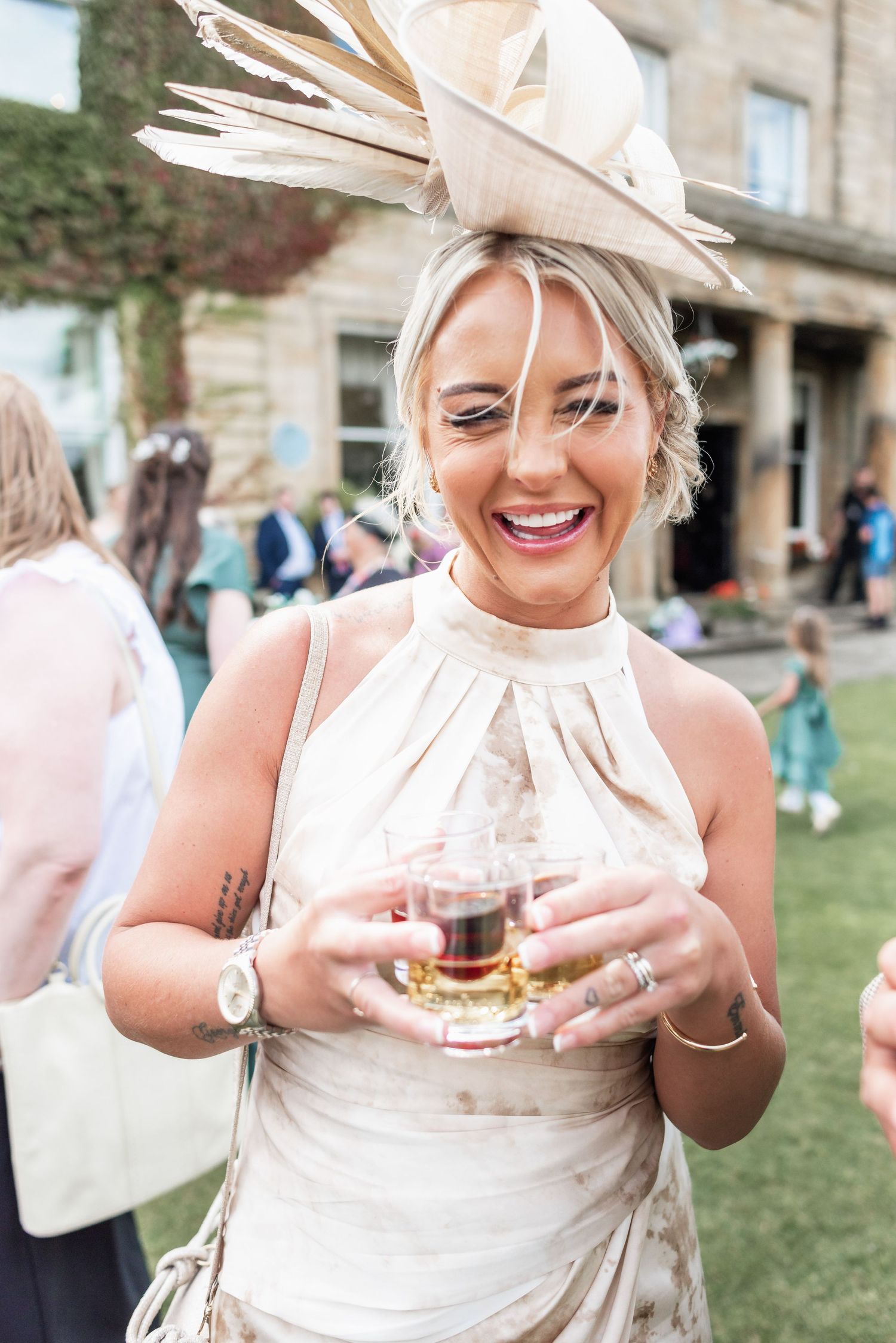 Wedding guest in cream dress and dramatic fascinator holds a drink while smiling at an outdoor reception.