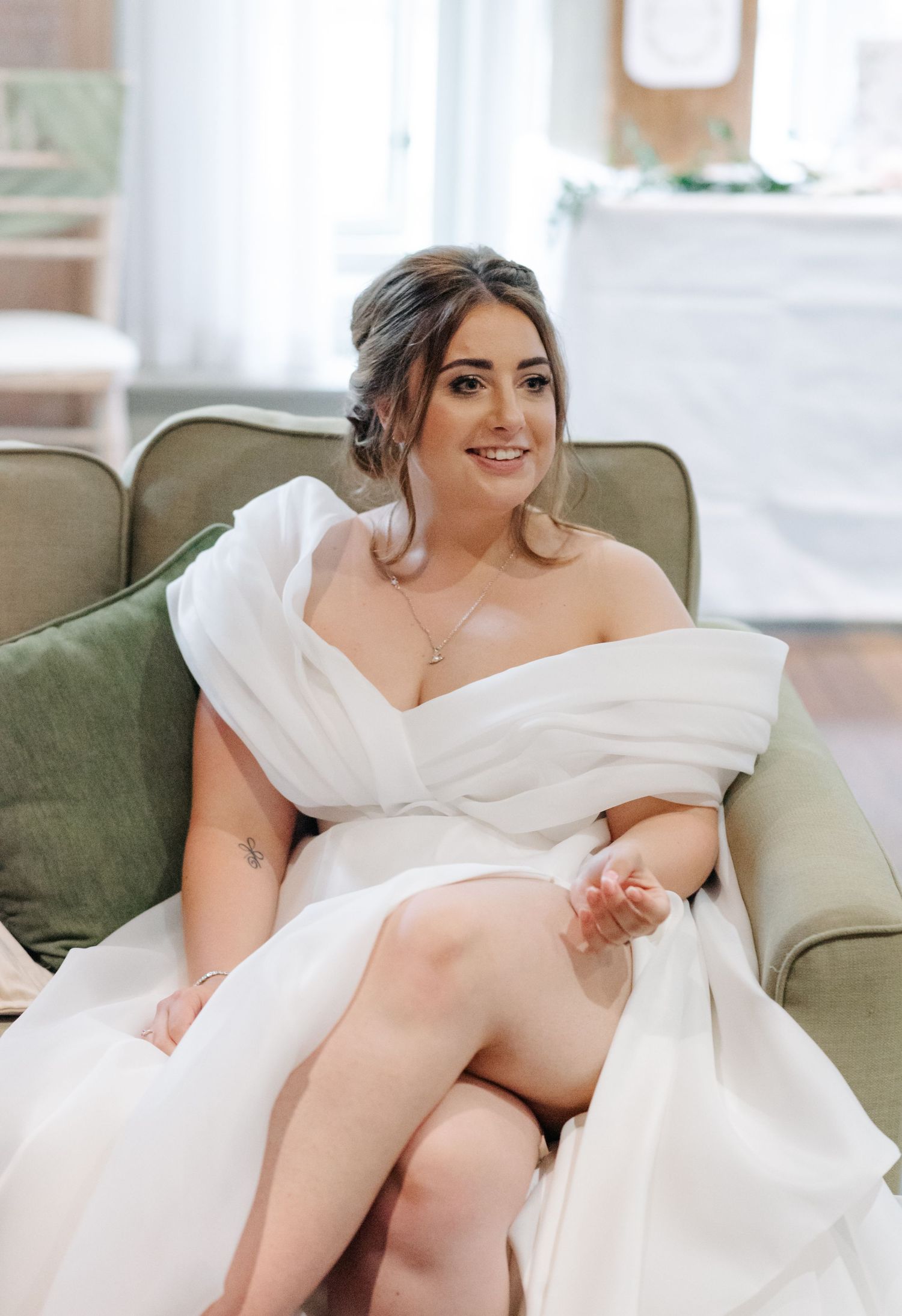 Bride in elegant white wedding dress lounges gracefully on sage green sofa in natural light.