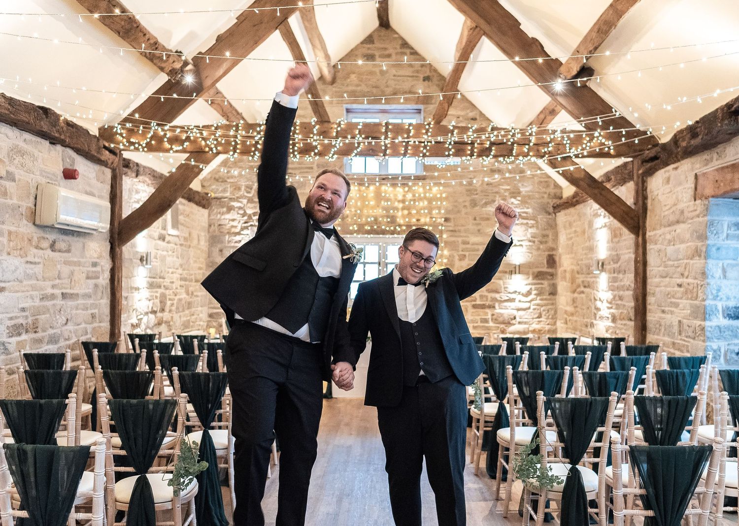Two people celebrating with raised arms in venue decorated with fairy lights and wooden beams.