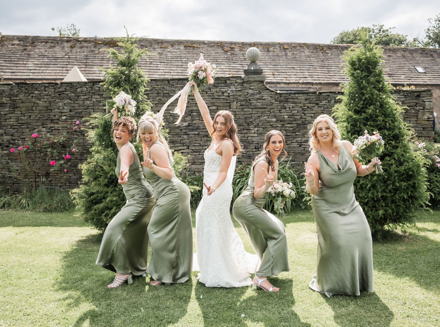 Bridal party in sage dresses celebrates joyfully in garden with raised bouquets.
