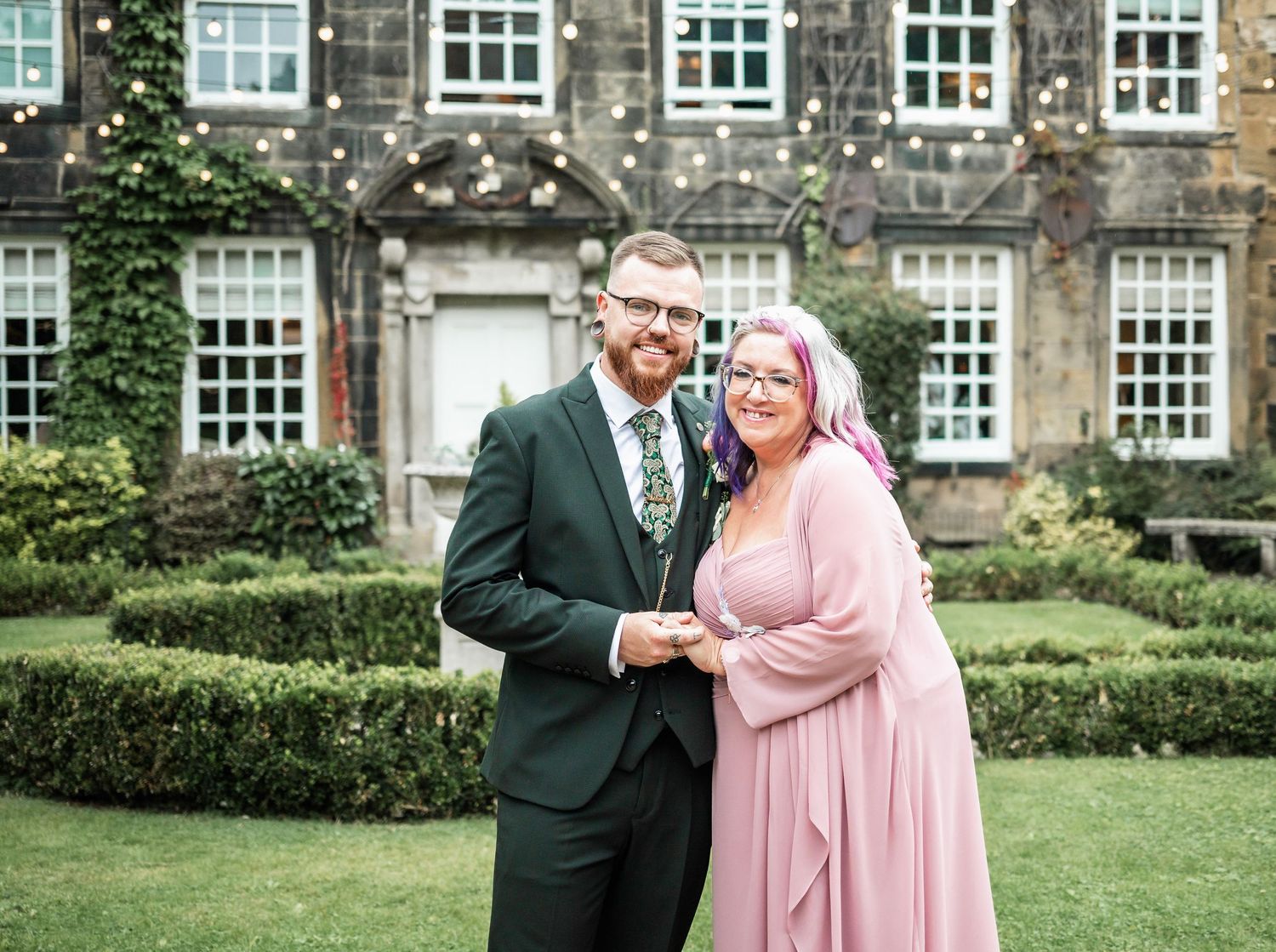 Couple poses together on the manicured lawn of a historic stone building with elegant windows and hedges.