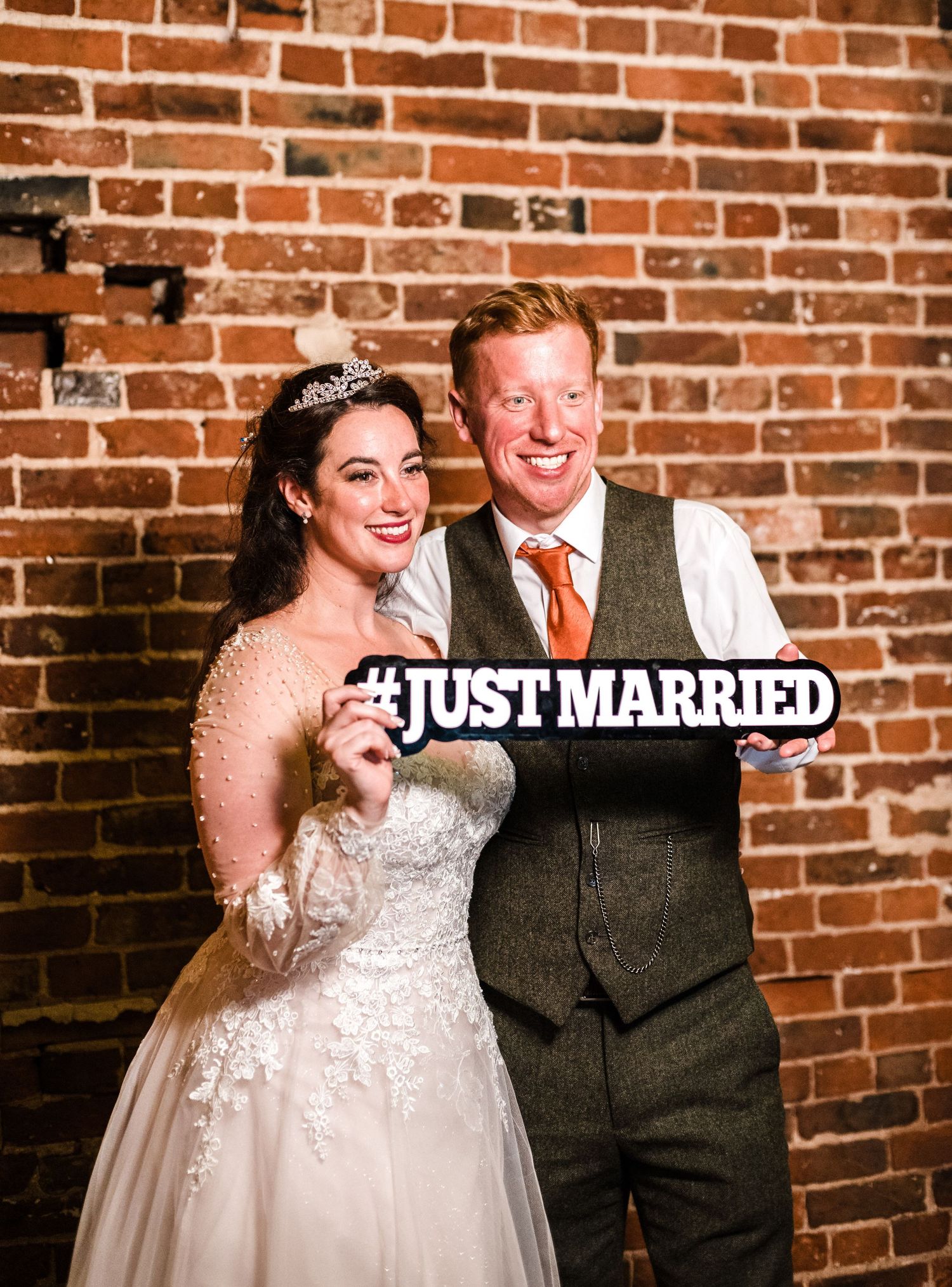 Newlyweds pose with a 'Just Married' sign against an exposed brick wall.