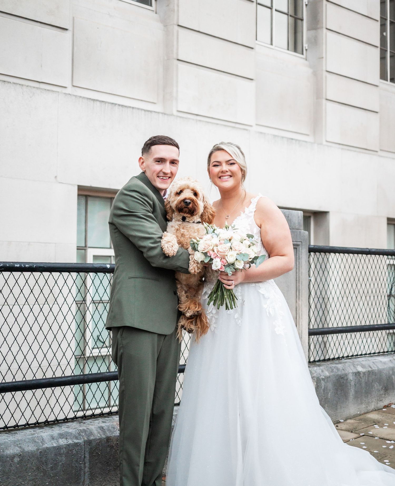 Wedding couple poses with their small brown dog while holding a pastel floral bouquet.