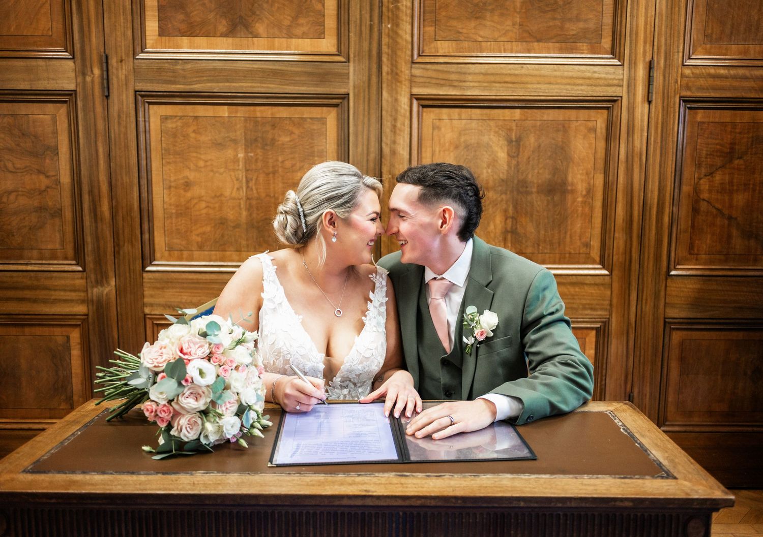 Wedding couple signs marriage documents at wooden desk in traditional venue.