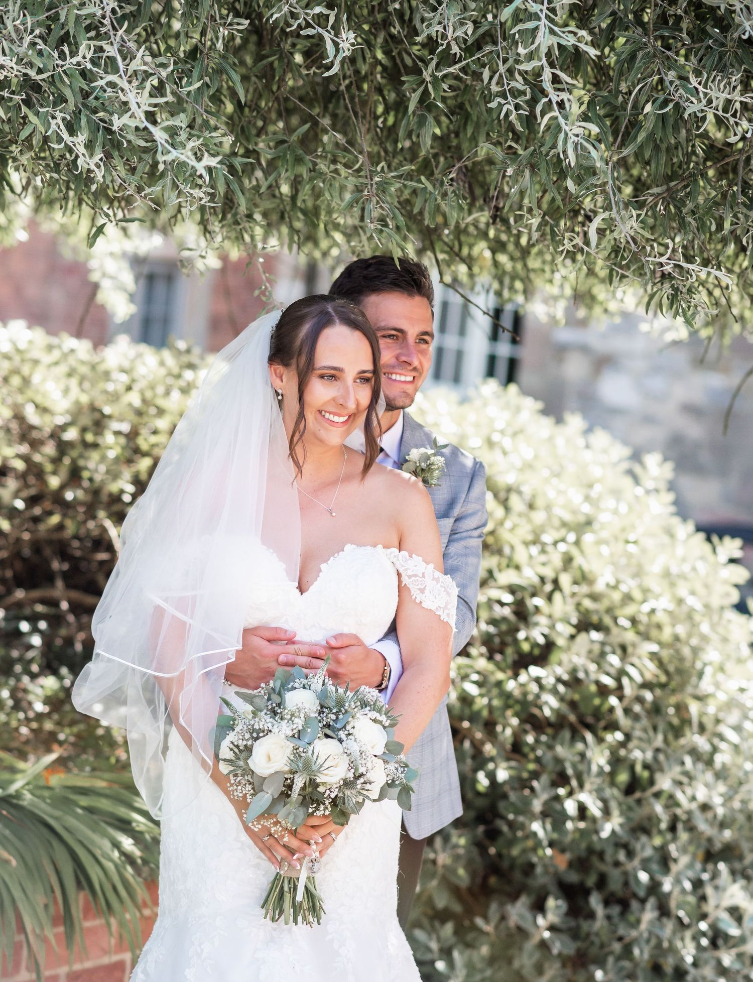 Newlywed couple poses under hanging greenery with bridal bouquet.
