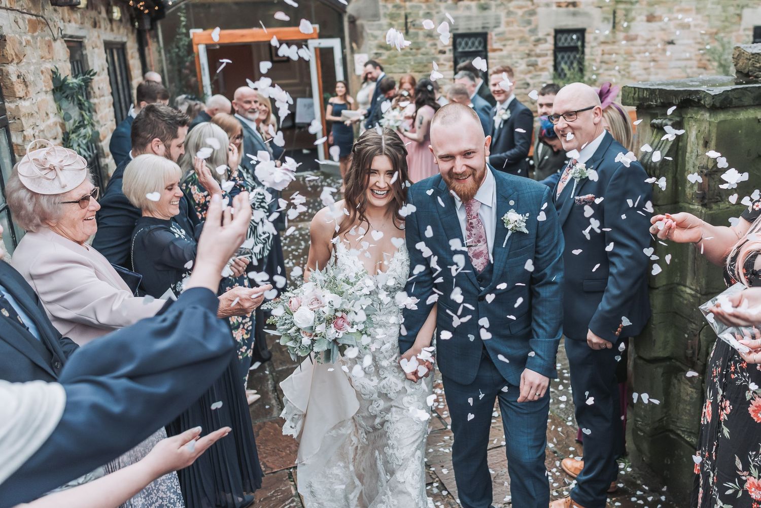 Wedding guests shower the newlyweds with confetti outside a stone building.