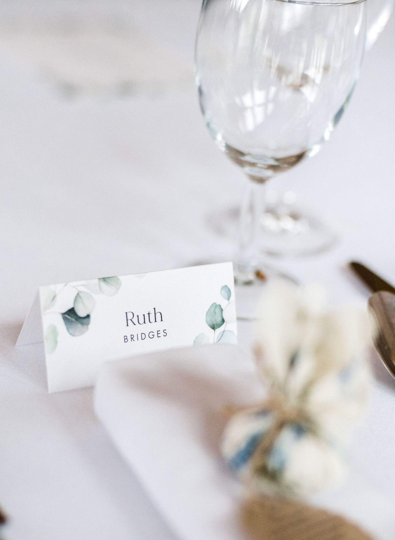Wedding place card with eucalyptus design next to wine glass on white tablecloth.