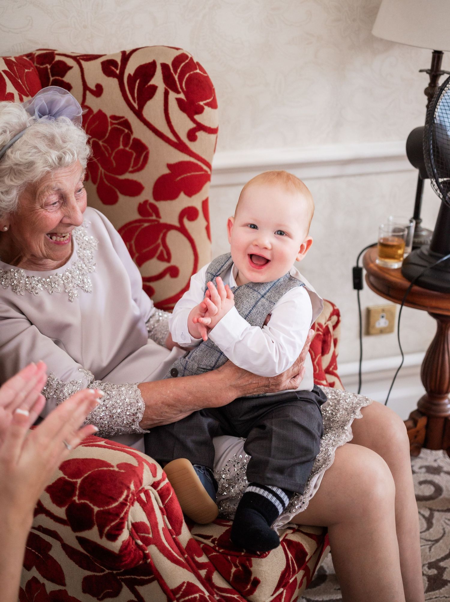 Happy baby in formal wear sits on lap in red floral chair during joyful moment.