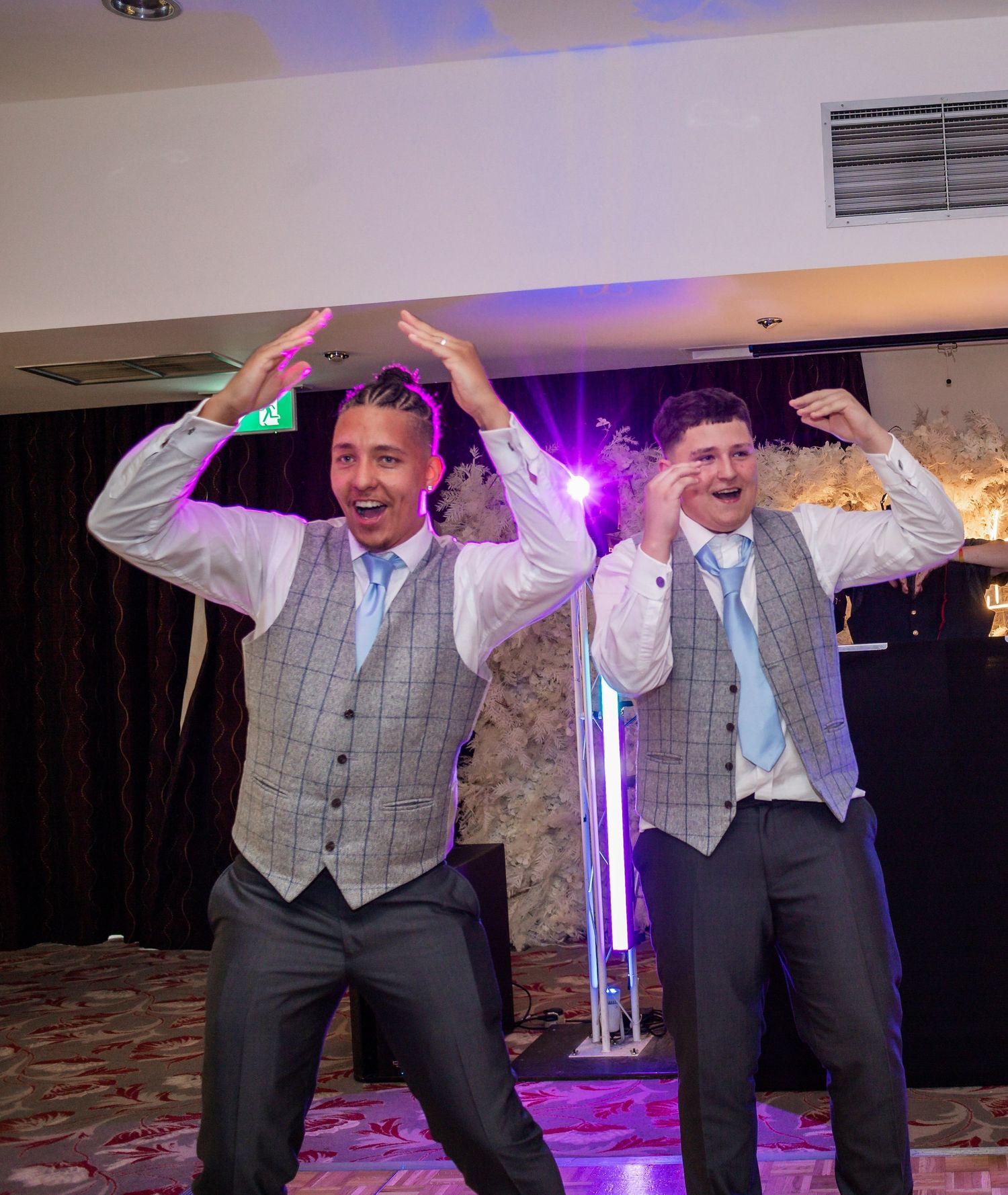 Two groomsmen in matching waistcoats dance energetically under purple lighting at a wedding reception.