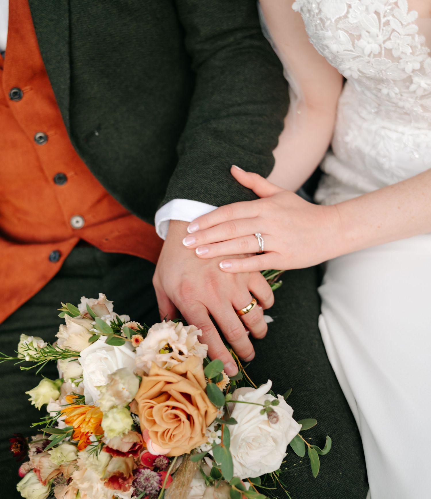 A bride and groom hold hands showing their wedding rings behind a rose bouquet, the groom wearing an orange waistcoat and tweed jacket.