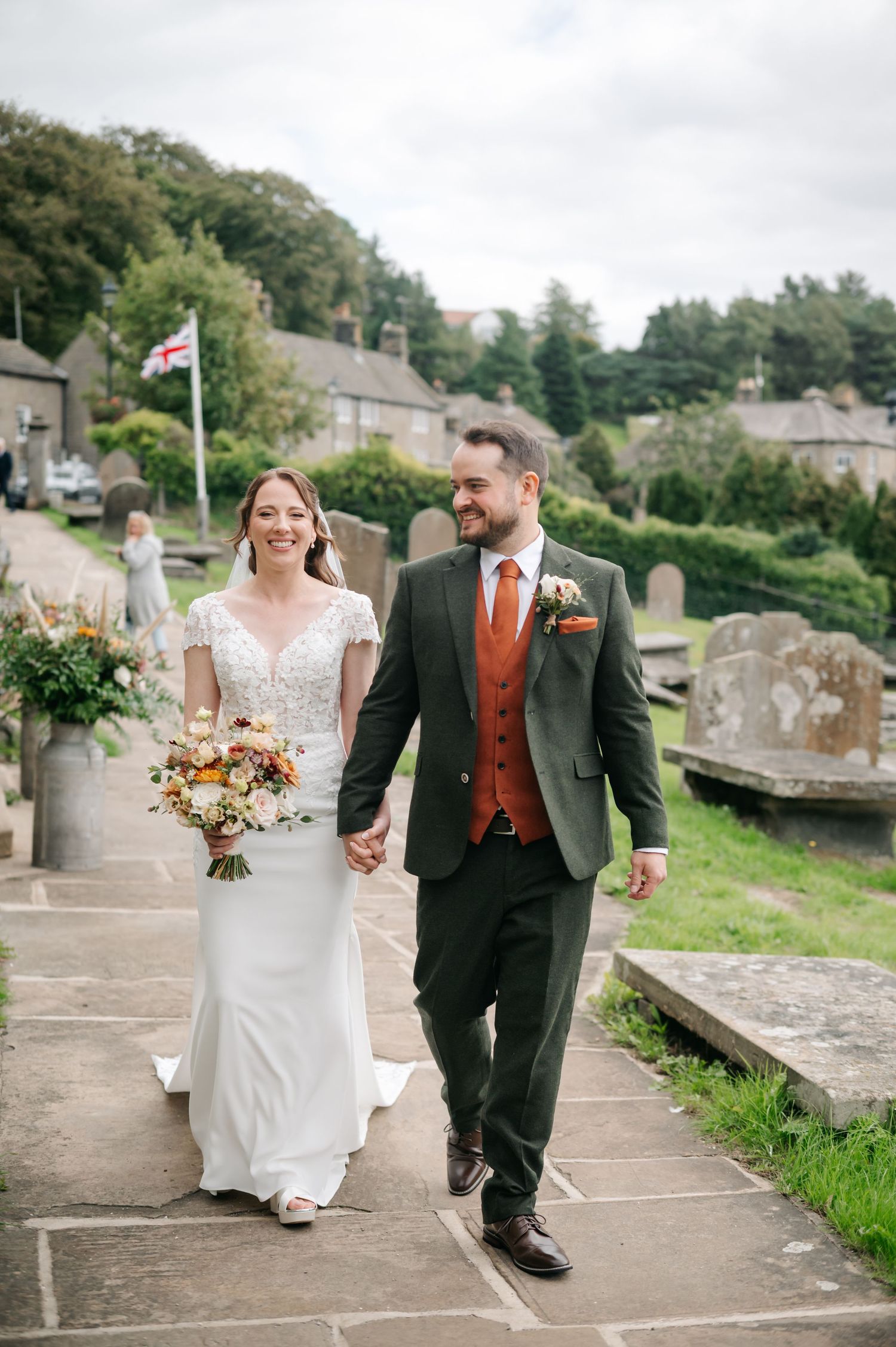 Smiling bride in a lace gown holding a bouquet walks hand in hand with a groom in a green tweed suit through a stone churchyard.