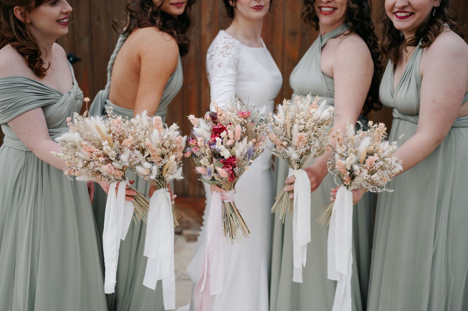 Bridesmaids in sage green dresses hold dried flower bouquets in a row.