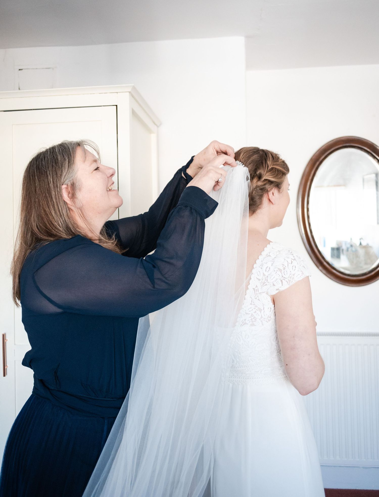 A person in navy blue adjusts a flowing white wedding veil on someone wearing a lace wedding dress.