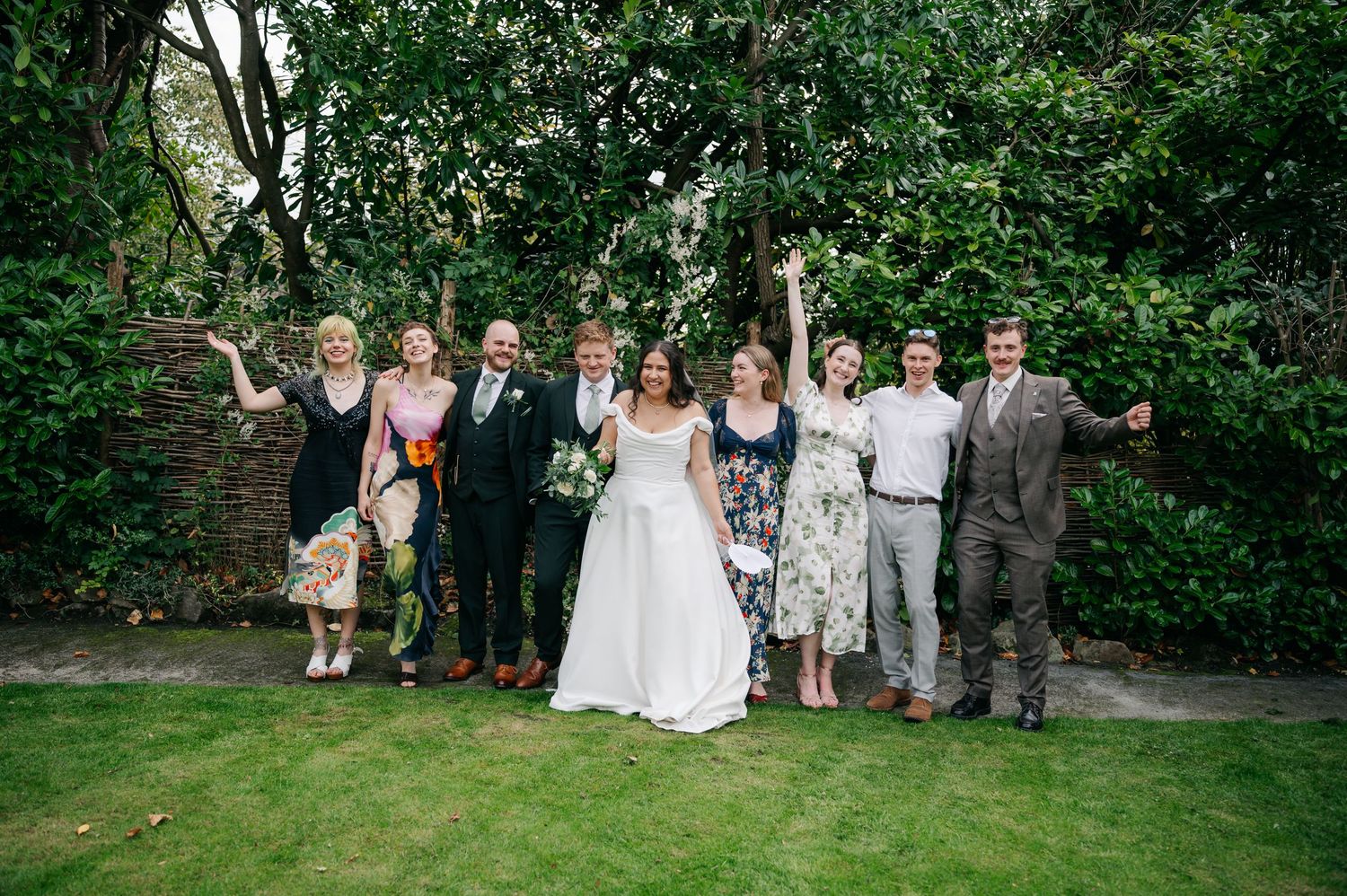 Wedding party and guests gathered outdoors for a group photo on a green lawn.