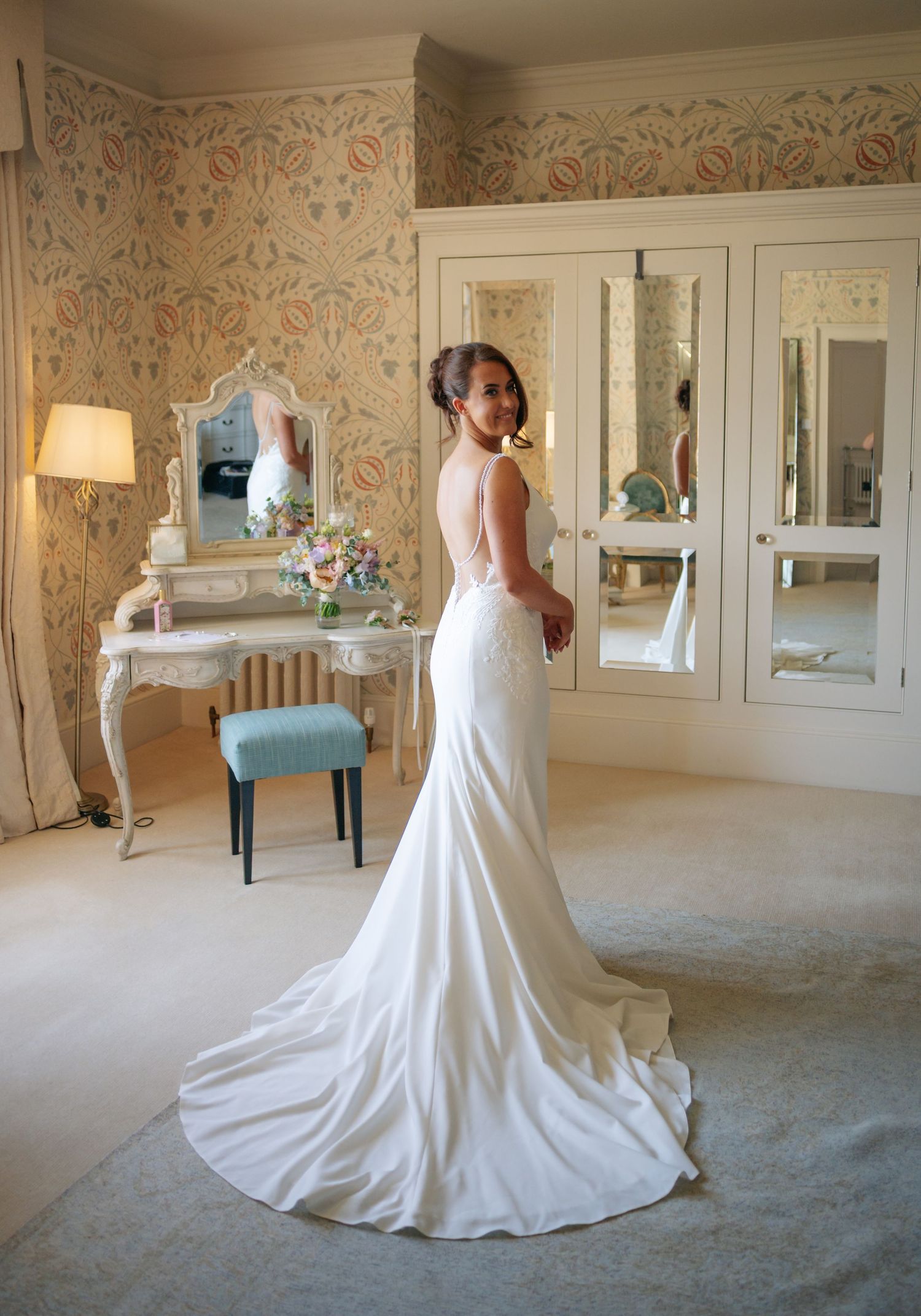 Elegant white wedding dress displayed in a room with ornate wallpaper and mirrors.