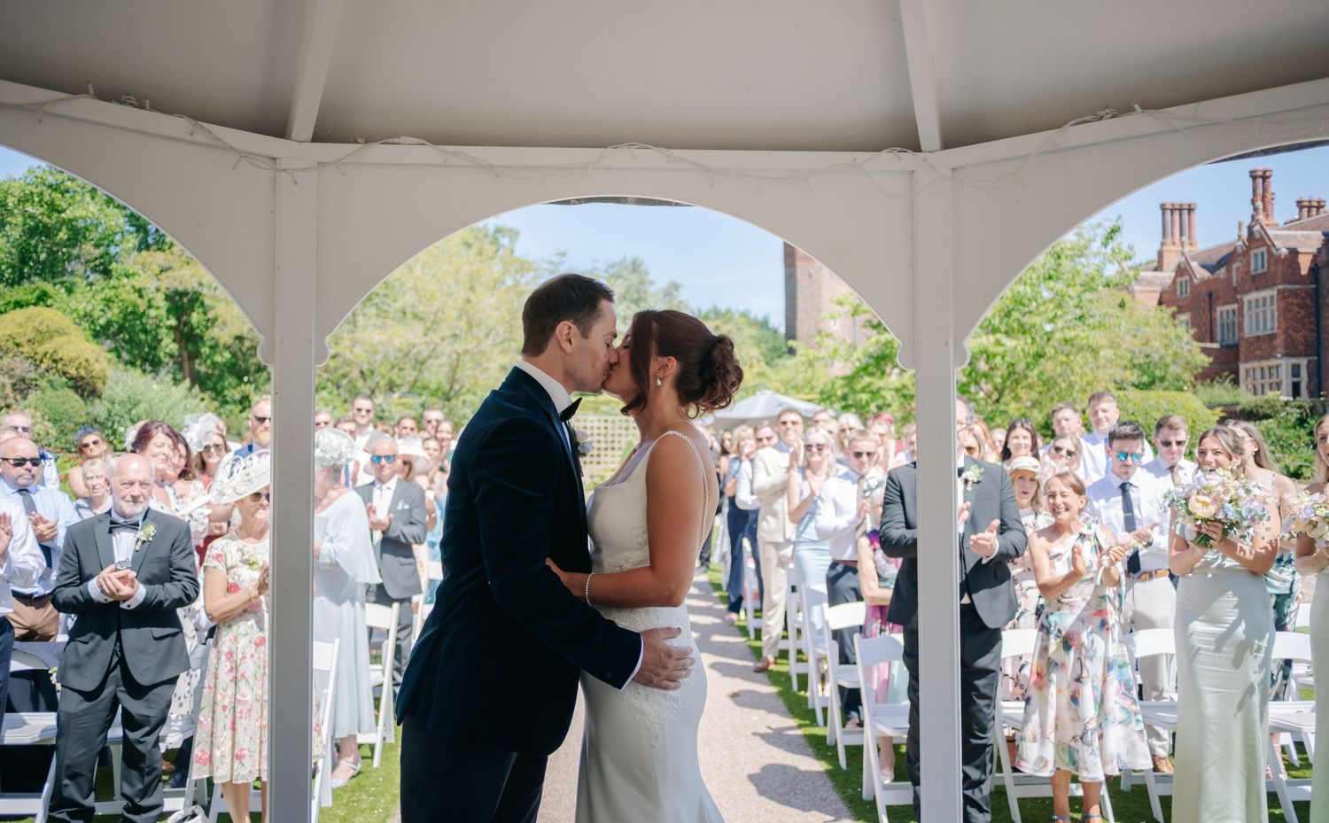 Wedding ceremony panorama showing guests gathered under a white tent on a sunny day.