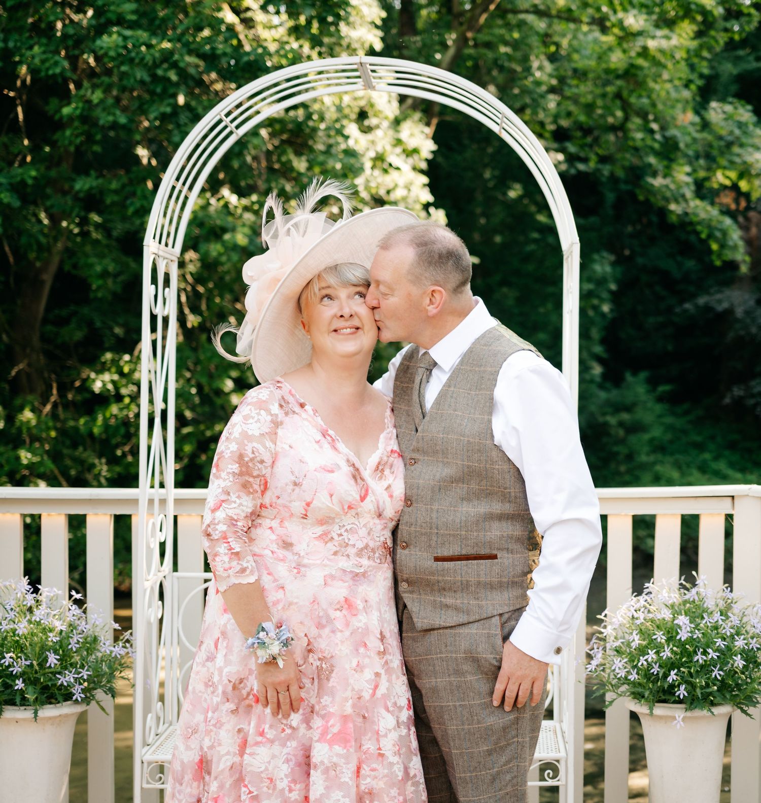 Couple sharing a kiss under a white garden arch, woman wearing a floral pink dress.