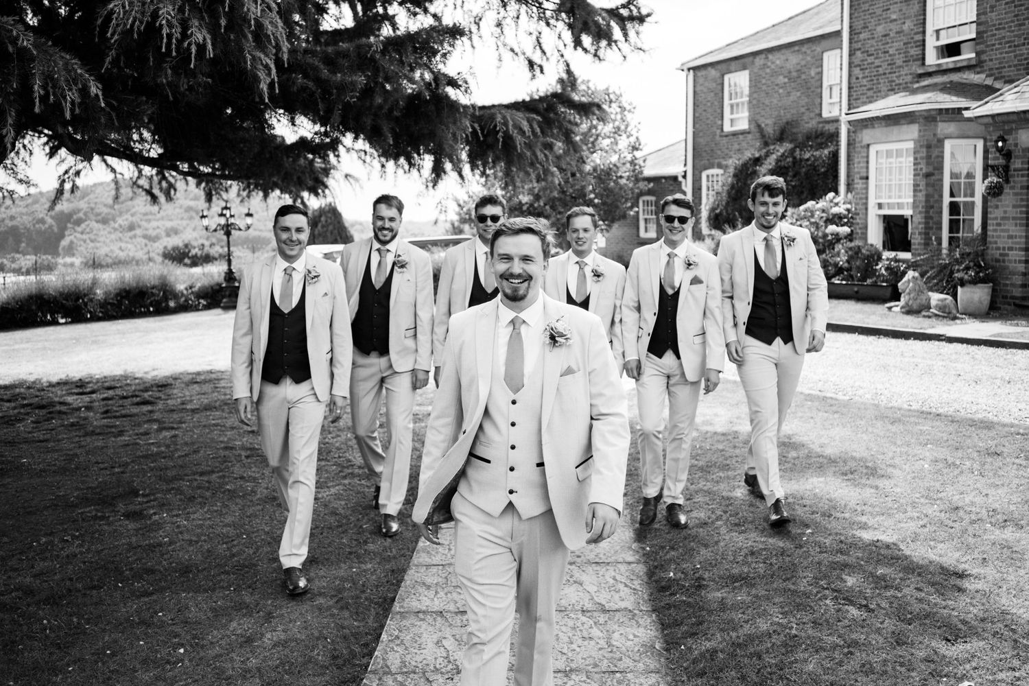 Groomsmen in light colored suits walk together down a path while laughing and smiling.