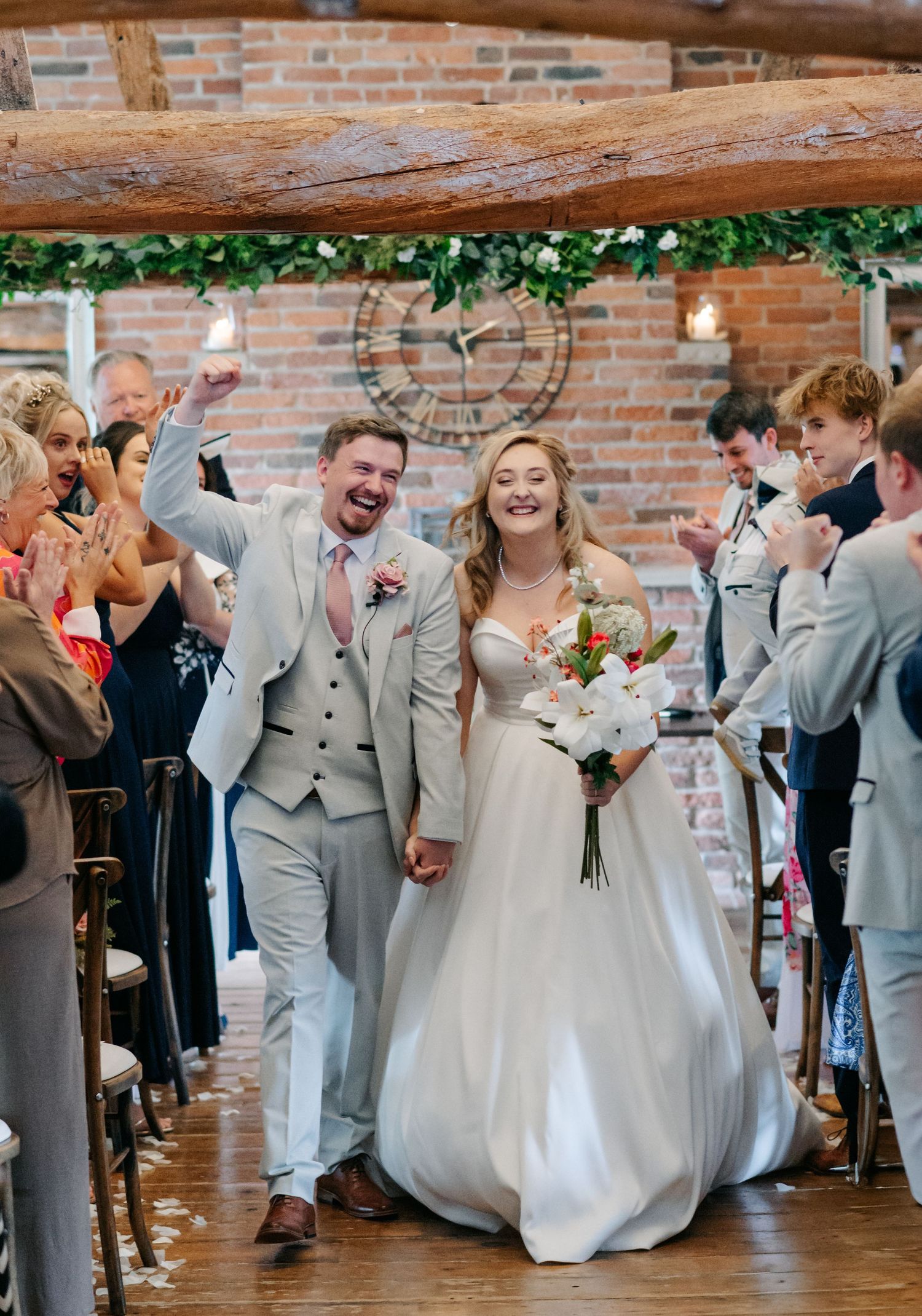 Wedding celebration with guests in gray suits and floral dresses dancing in a rustic indoor venue.