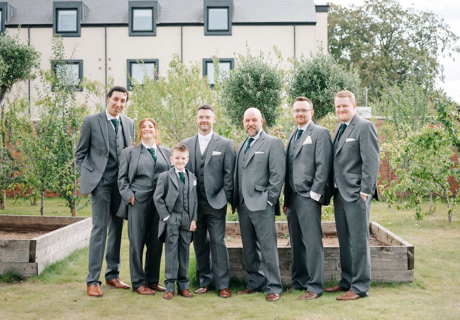A group of groomsmen in matching gray suits pose together in a garden setting.
