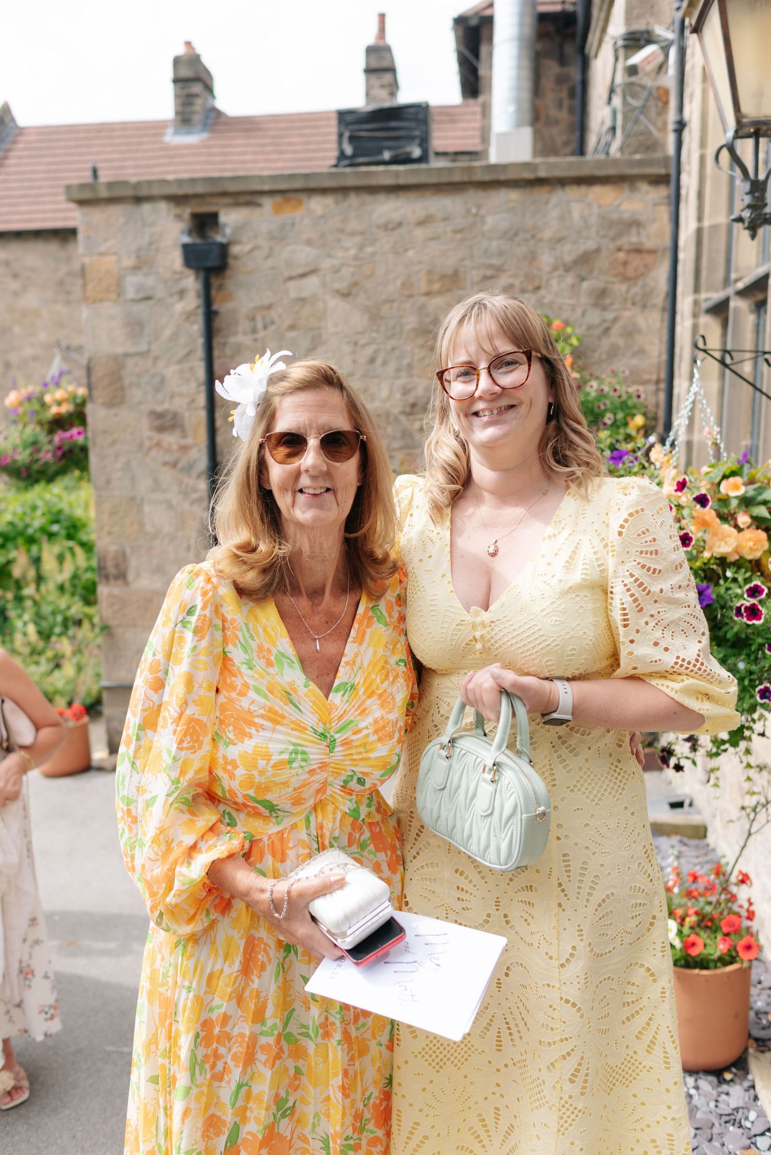 Cheerful wedding guests in bright summer dresses pose against stone building backdrop.