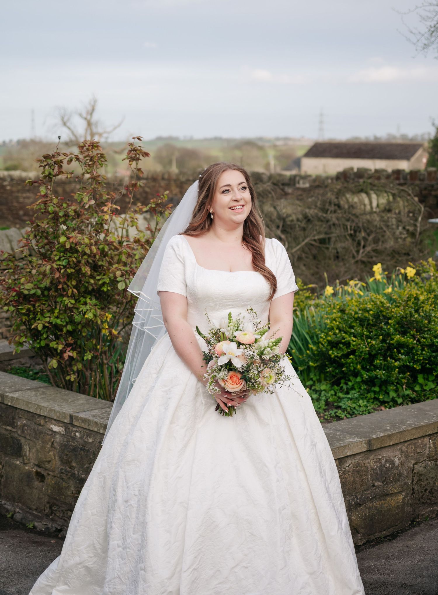 A bride in a white wedding dress with short sleeves and a veil holds a pastel bouquet while standing in a garden setting.