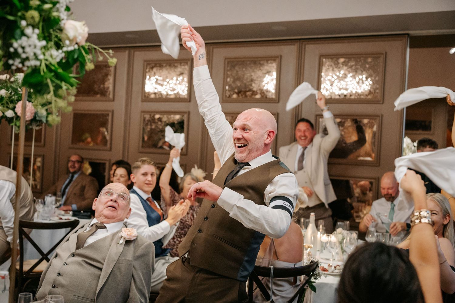 Wedding reception guests waving napkins in celebration at elegant venue with framed artwork.