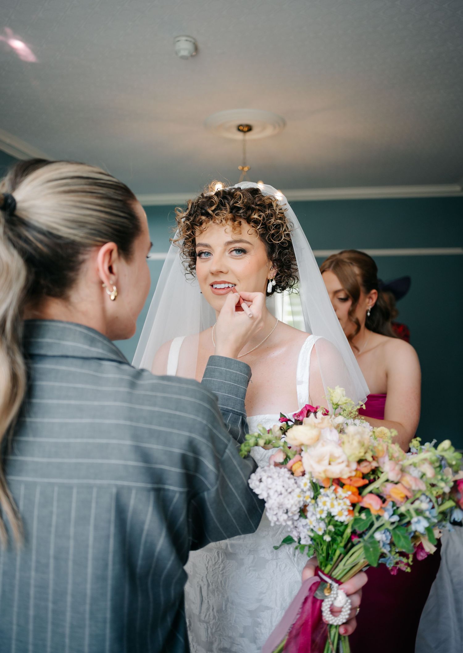 A touching moment during wedding preparations as final makeup touches are applied to the bride holding a floral bouquet.