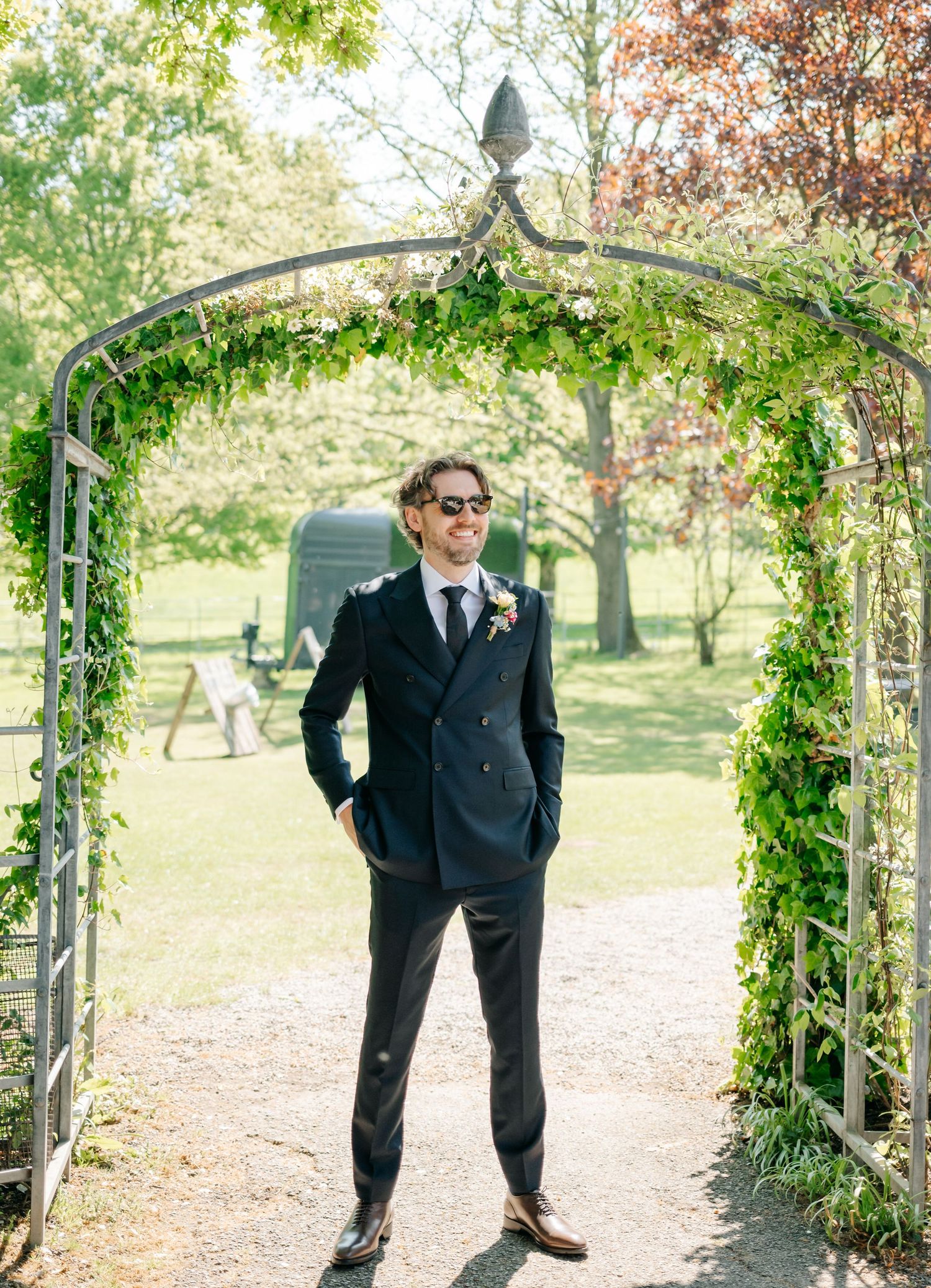 Wedding party member poses playfully under a vine-covered garden arch in a navy suit.
