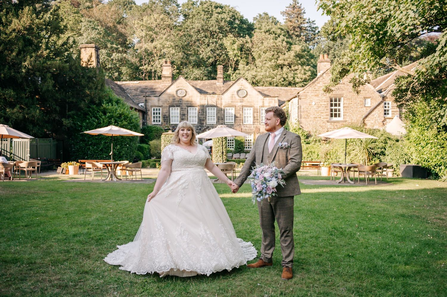 Bride and groom holding hands on a sunny lawn in front of a stone country house.