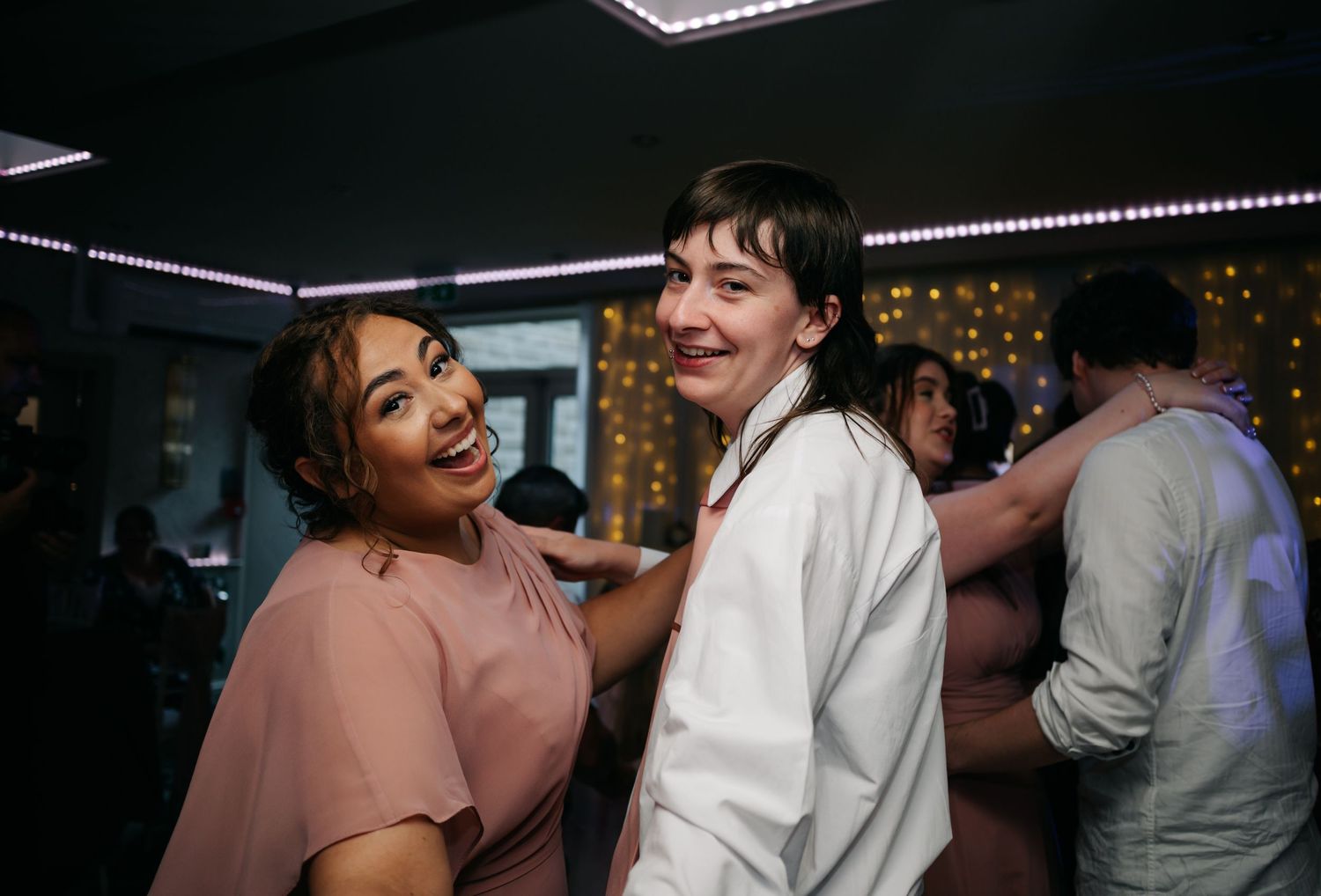 Two wedding guests smiling and dancing together at a lively indoor wedding reception celebration.