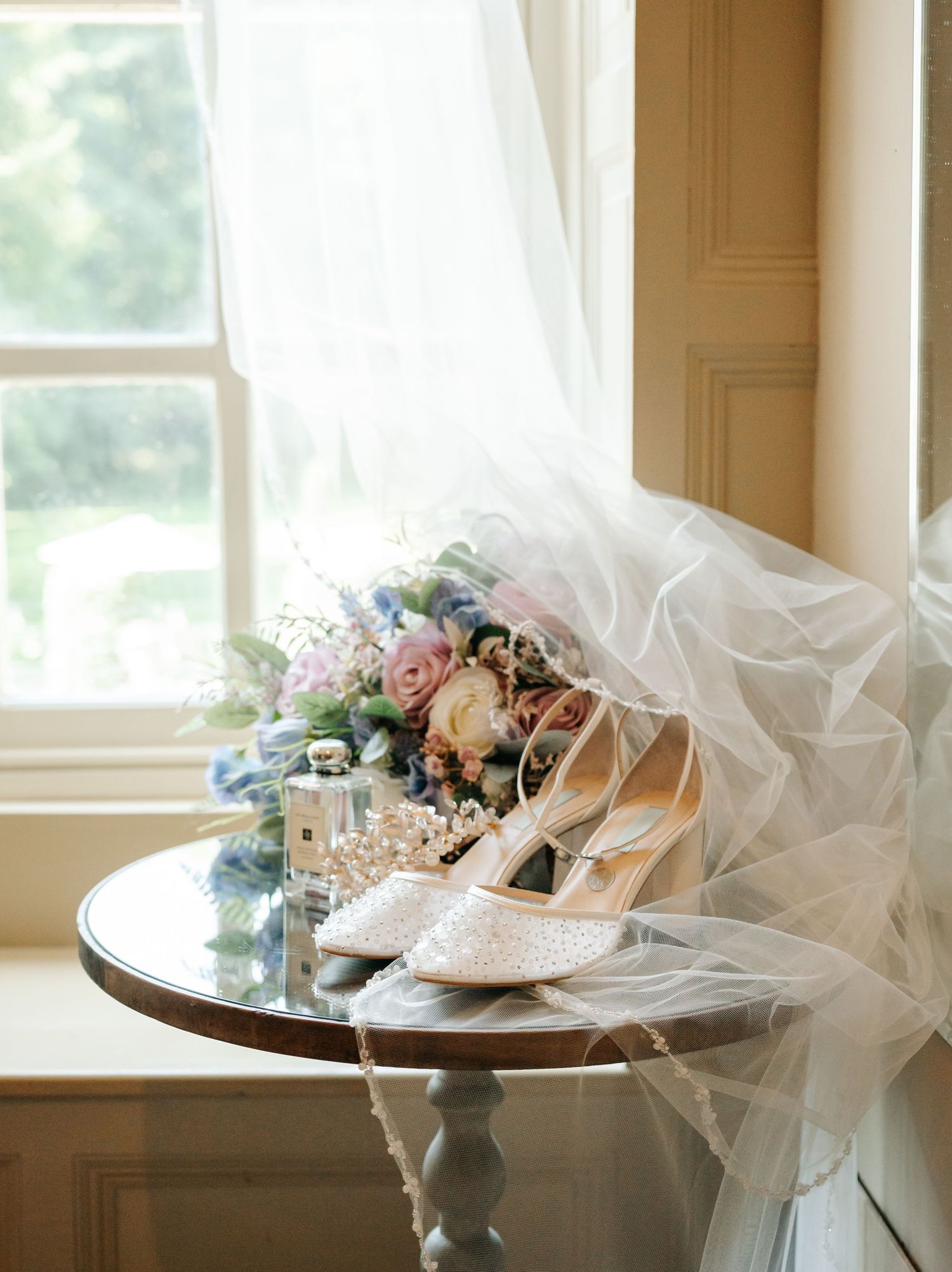 Bridal details flat lay with pearl heels, floral bouquet, veil, and perfume on a wooden table by a window.